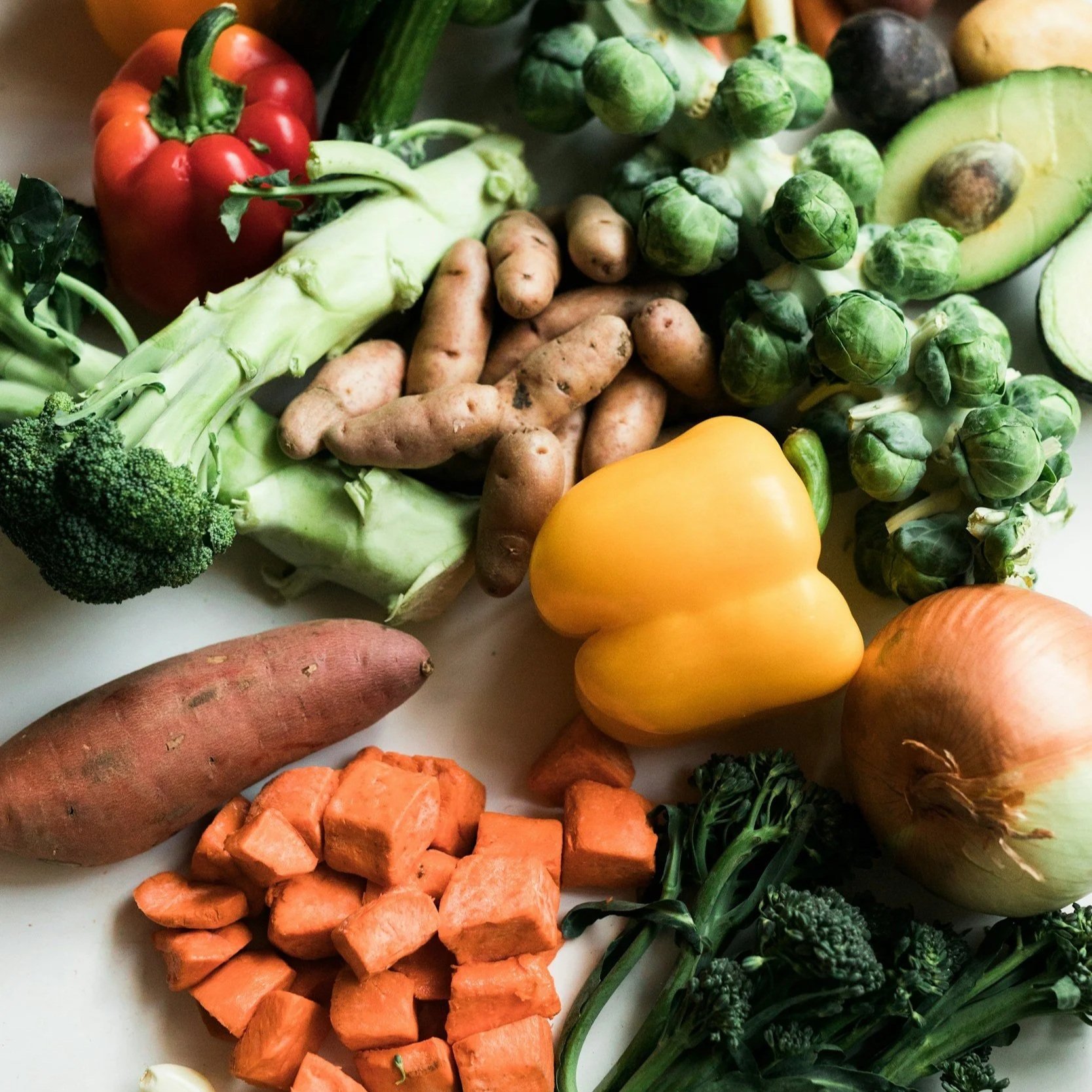 Assortment of fresh vegetables including red bell pepper, broccoli, sweet potatoes, zucchini, Brussels sprouts, avocado, yellow bell pepper, carrots, and broccoli.