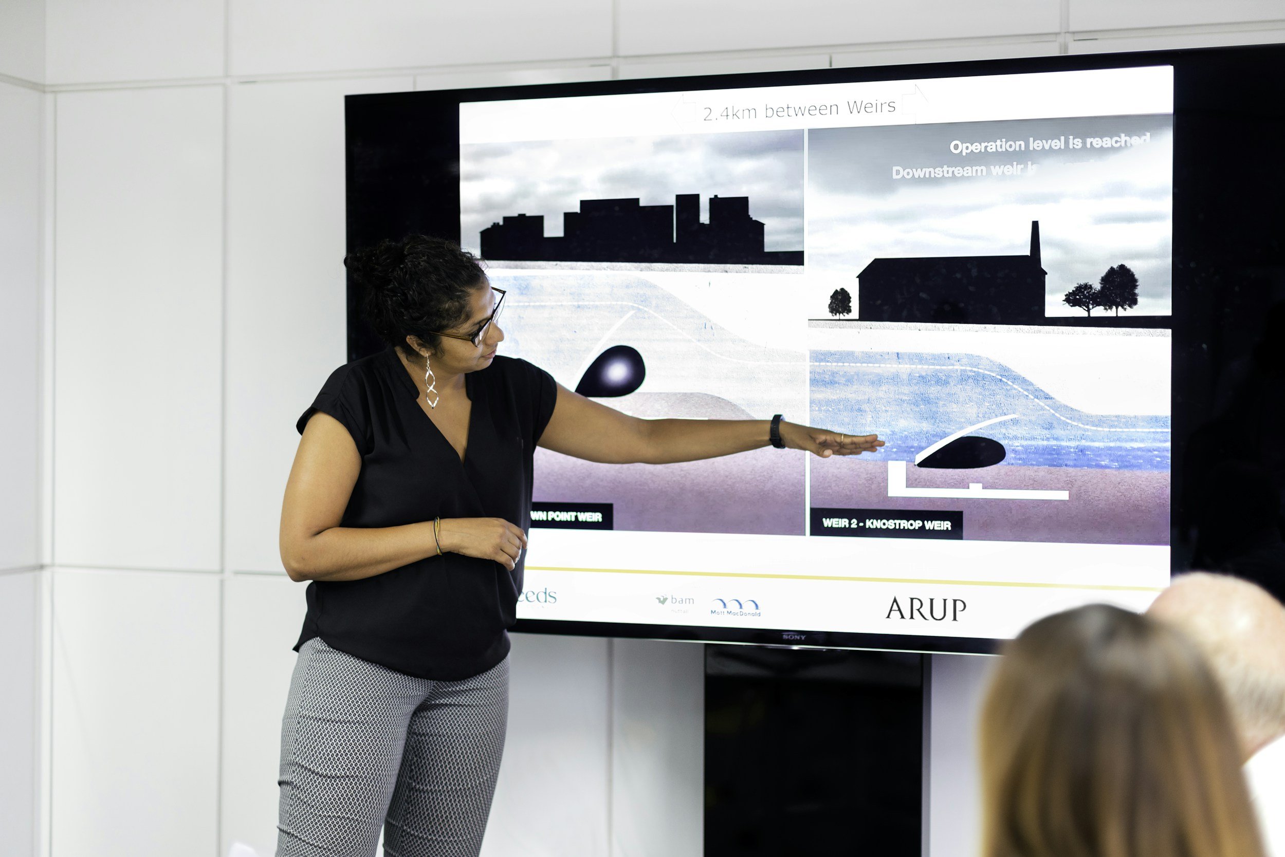 A woman with curly hair, glasses, and earrings is giving a presentation in front of a large screen displaying diagrams and text about weirs and water management.