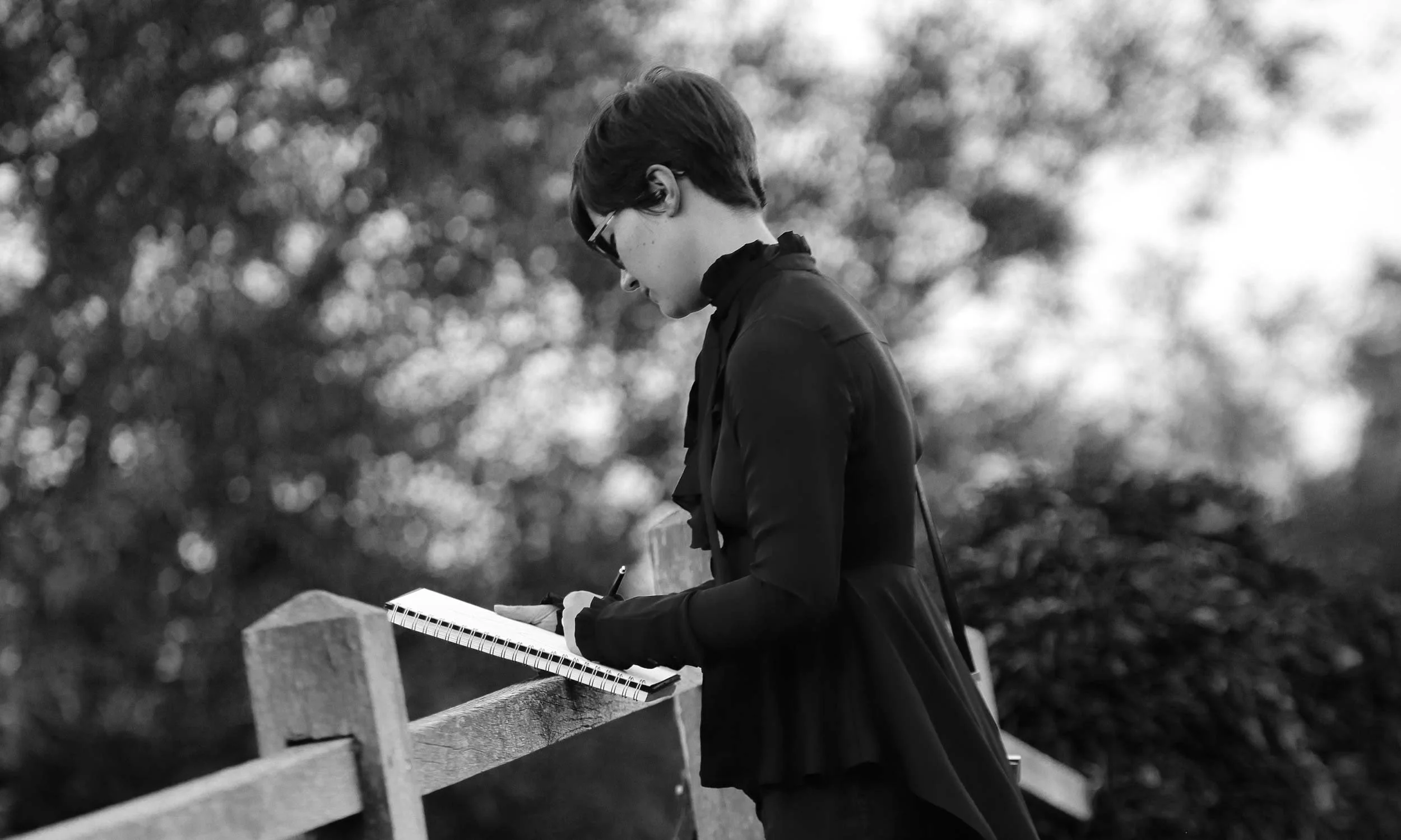 A woman with short hair and glasses sitting on a park bench, reading a book with a notebook inside, outdoors with blurred trees in the background.