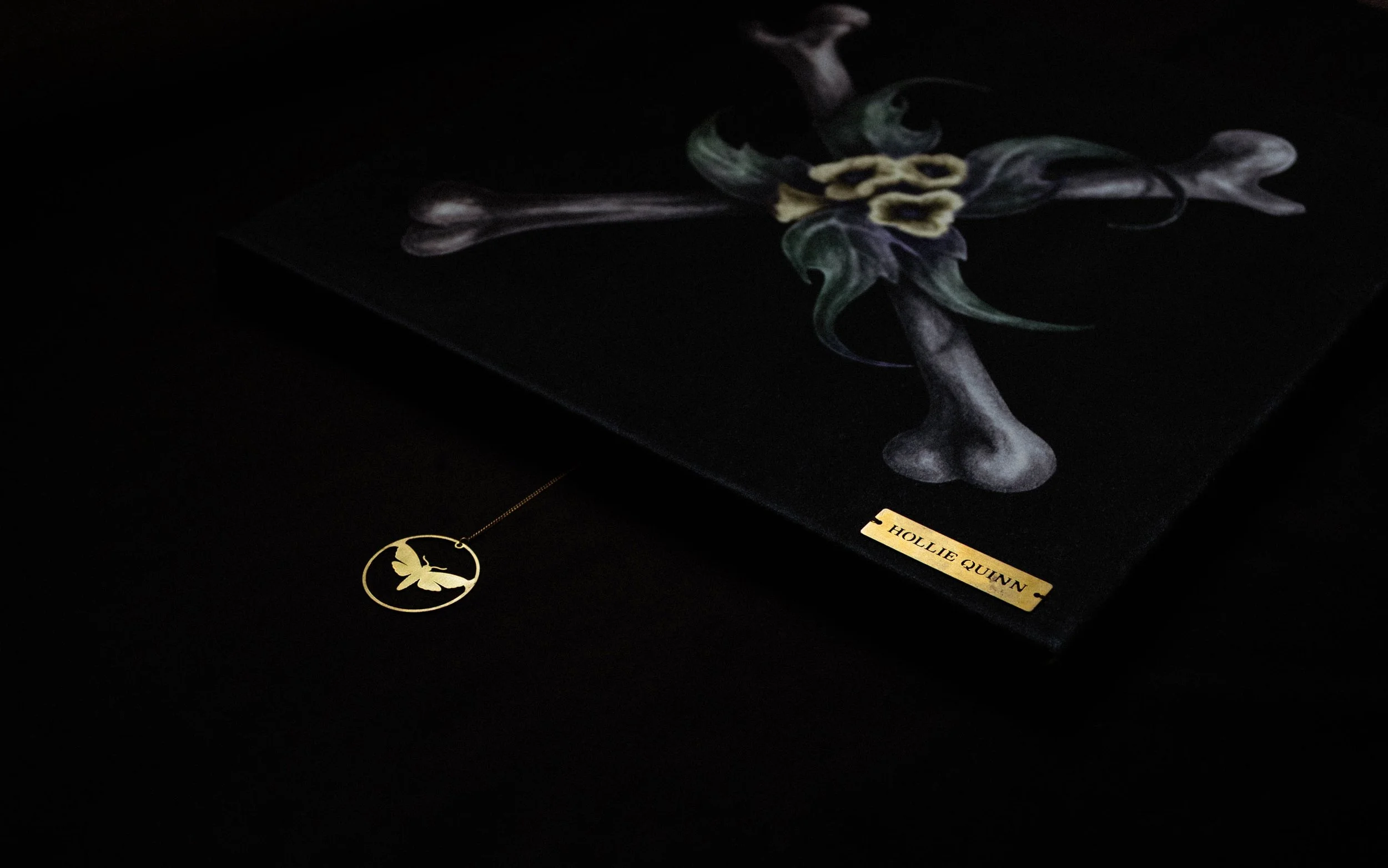 A dark display case with a black background showcasing an artwork by Hollie Quinn, featuring a colorful abstract design. A golden necklace with a hummingbird pendant is placed in front of the artwork, and a small label with the artist's name is visible.
