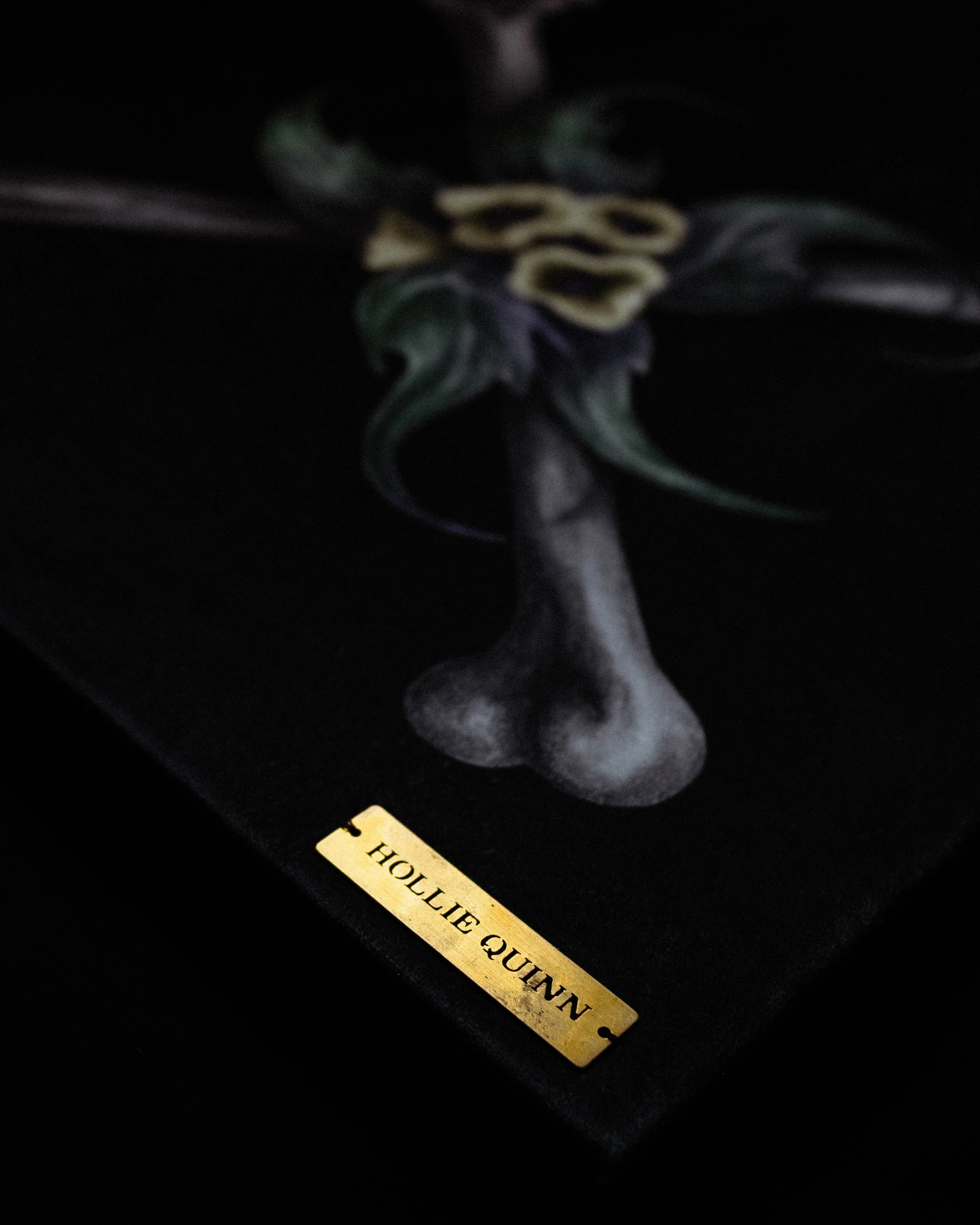 A dark background with a golden label reading 'HOLLIE QUAINT' and a black and yellow ribbon partially visible.