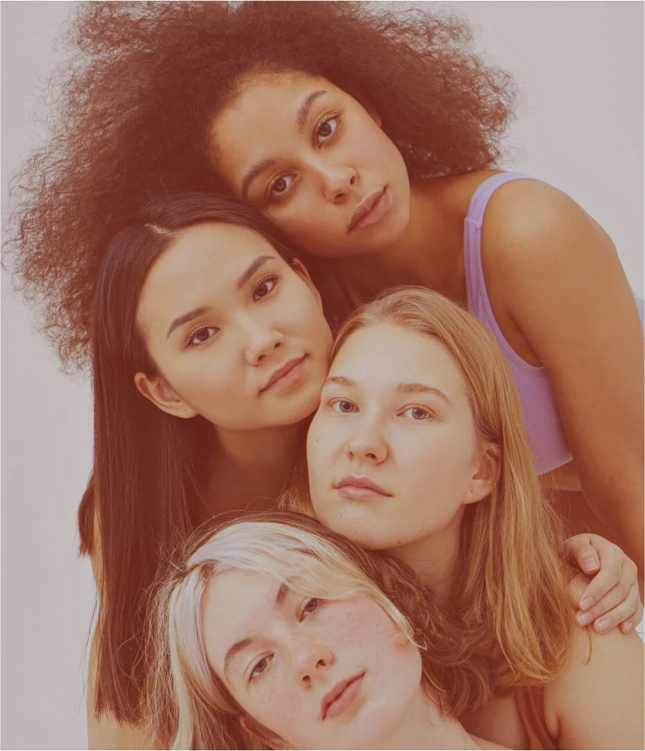 Cinco mulheres jovens de diferentes etnias posando juntas, uma apoiada na outra, olhando para a câmera.