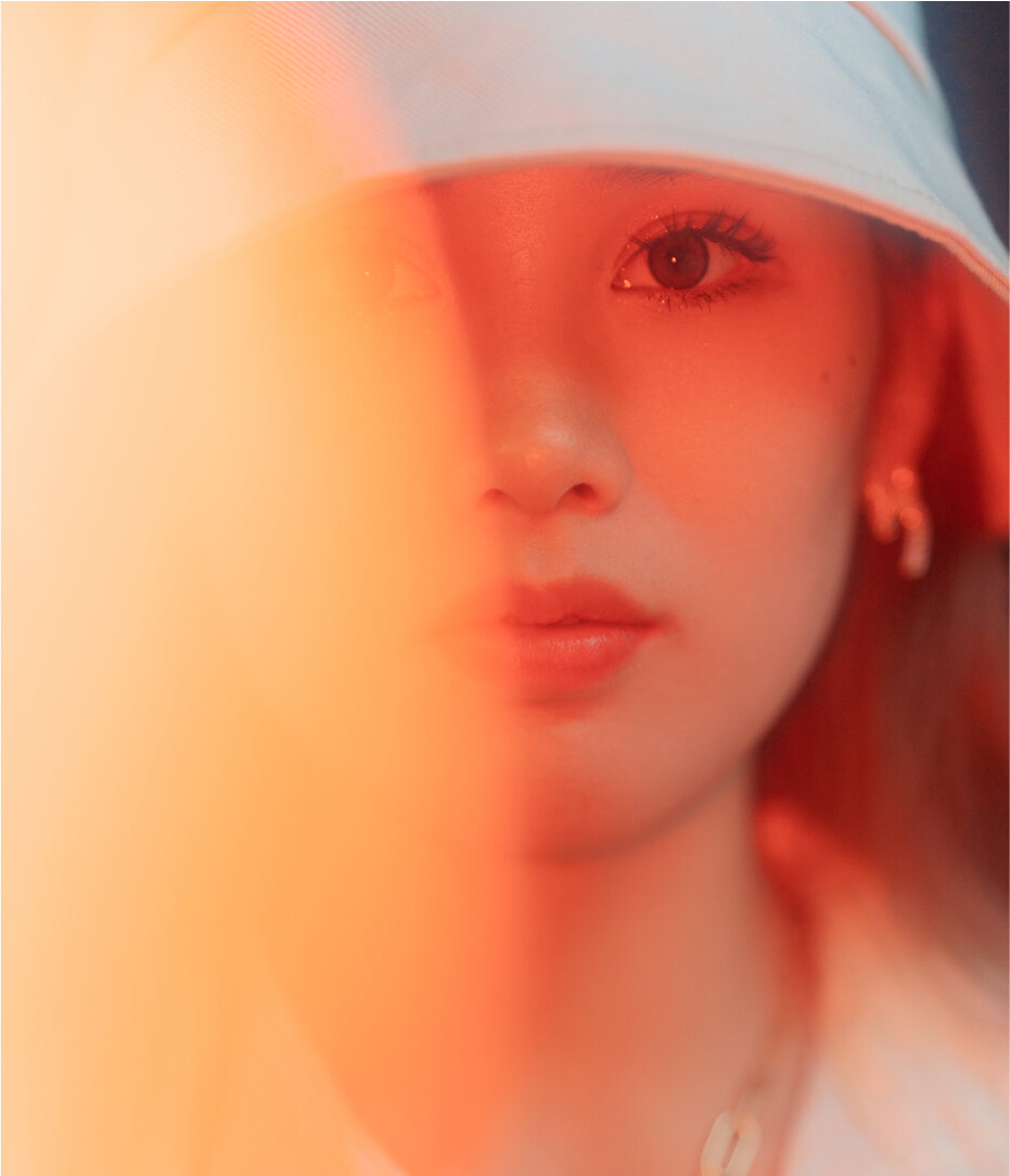 Retrato de uma jovem mulher com cabelo castanho-avermelhado e pele clara, usando brincos de argola e uma blusa de capuz, com uma iluminação com tons quentes e um efeito de desfoque na parte esquerda da imagem.