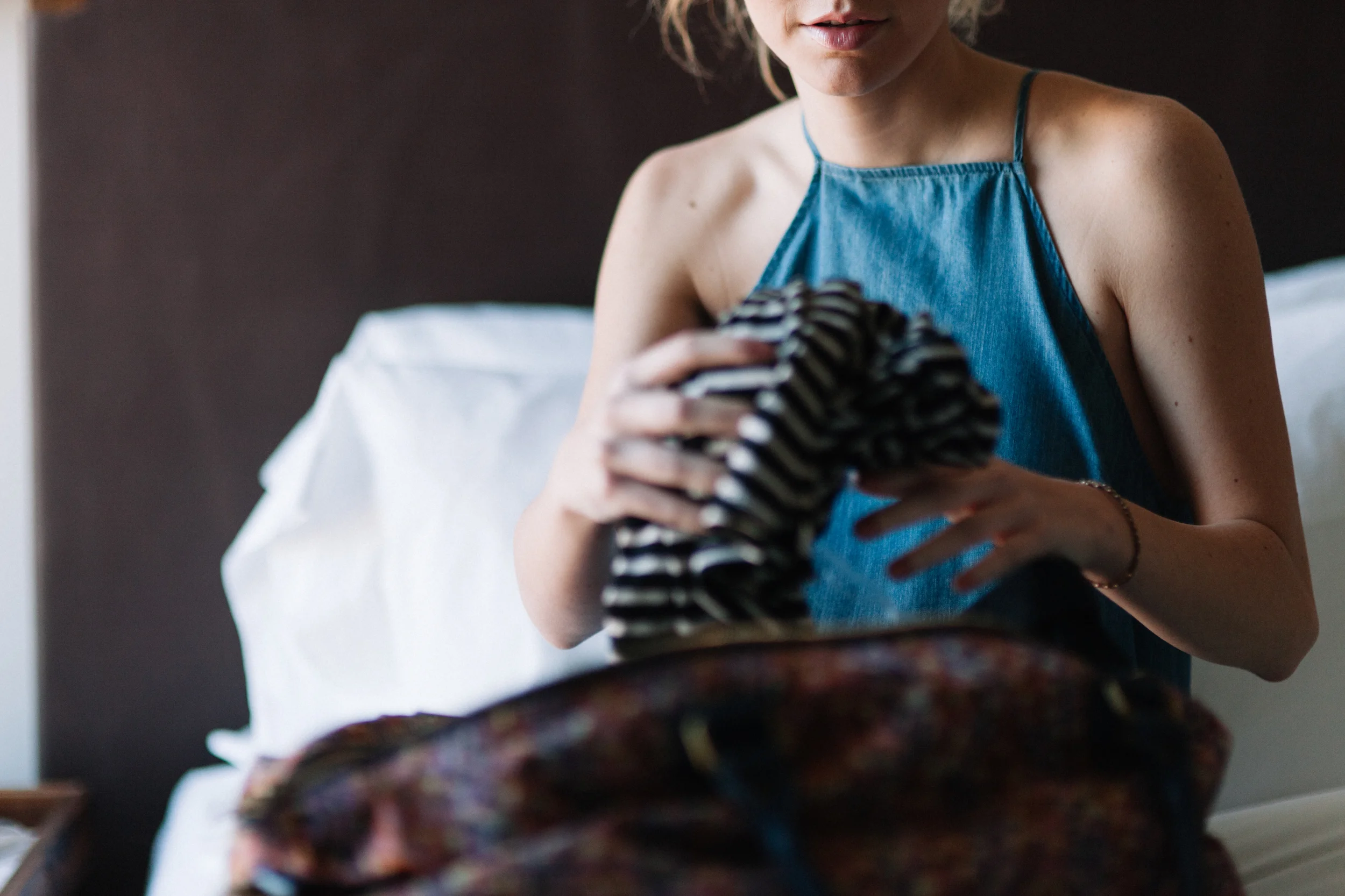 Mulher sentada na cama, segurando uma roupa deitada sobre as pernas, usando vestido azul de alças finas