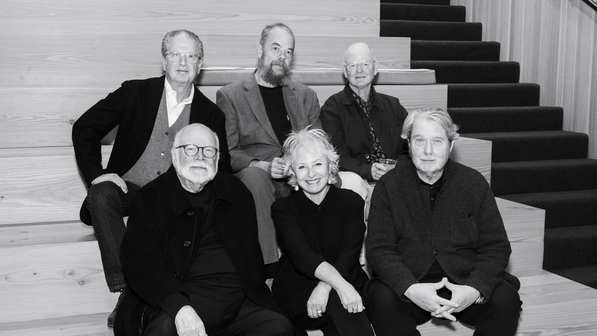 A group of seven diverse people, four men and three women, gathered together on a staircase with wooden steps. They are smiling and looking at the camera, dressed in casual to semi-formal clothing, in a modern indoor setting.