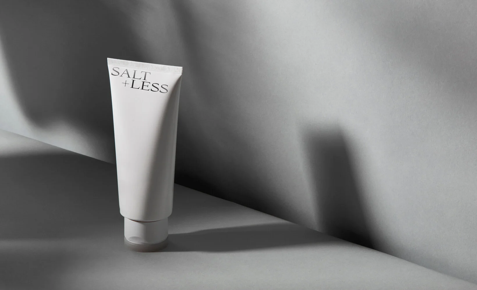 Tubo de creme branco com a frase "Salt + Less" escrito na parte superior, posicionando-se de pé sobre uma superfície cinza, com sombra projetada ao fundo.