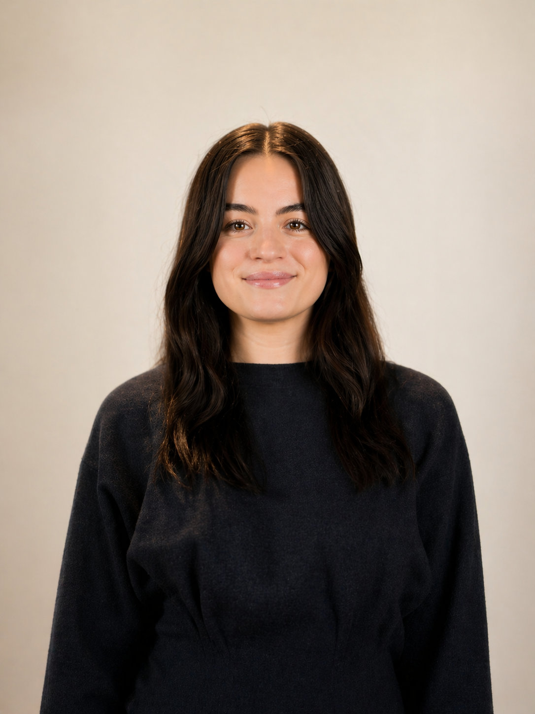 A young woman with long dark brown hair, wearing a black long-sleeved shirt, smiling in front of a plain off-white background.