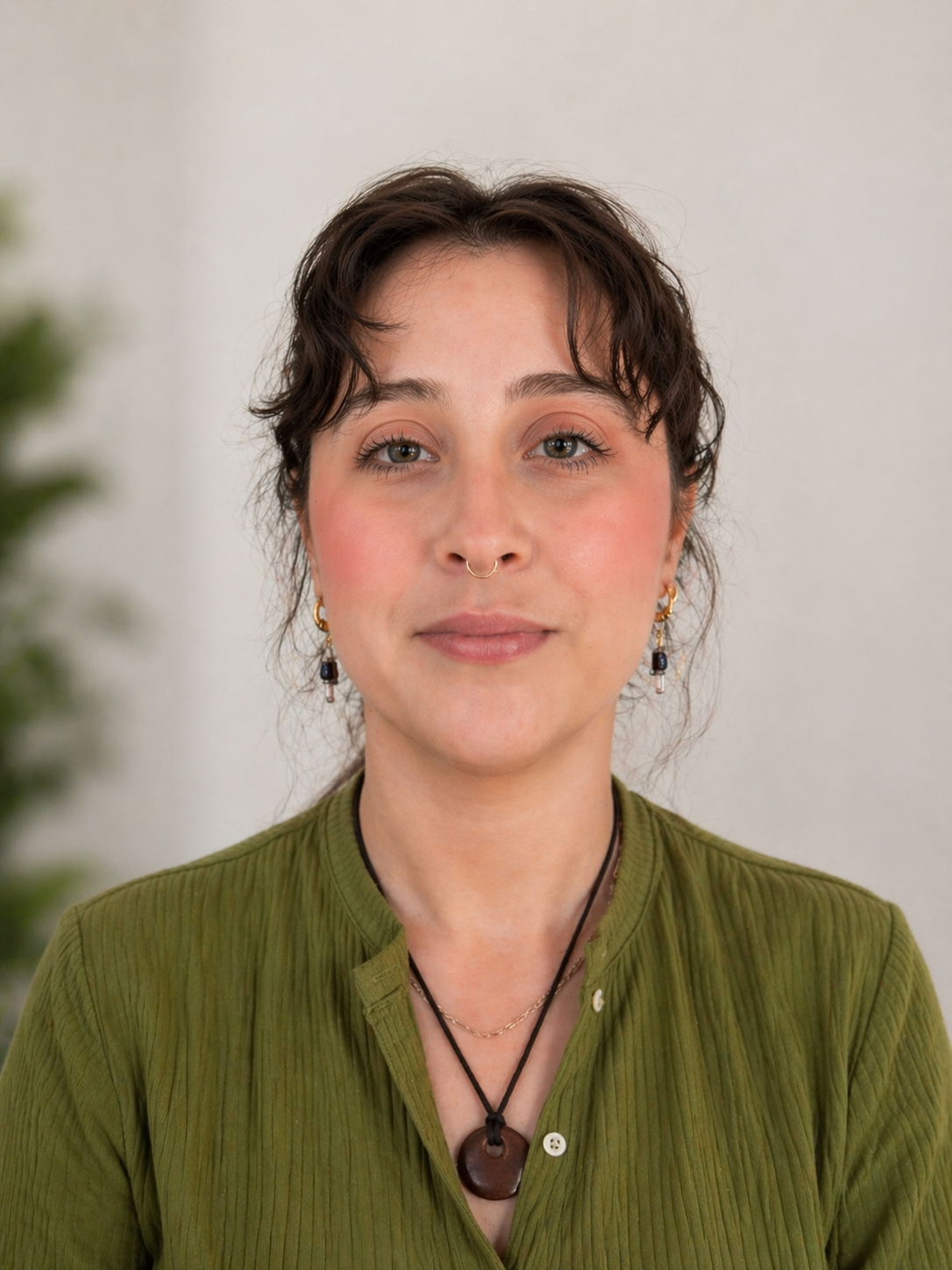 A person with short brown hair, light skin, and blue eyes, wearing an olive green shirt, layered necklaces, earrings, and a septum ring, standing against a plain white wall.