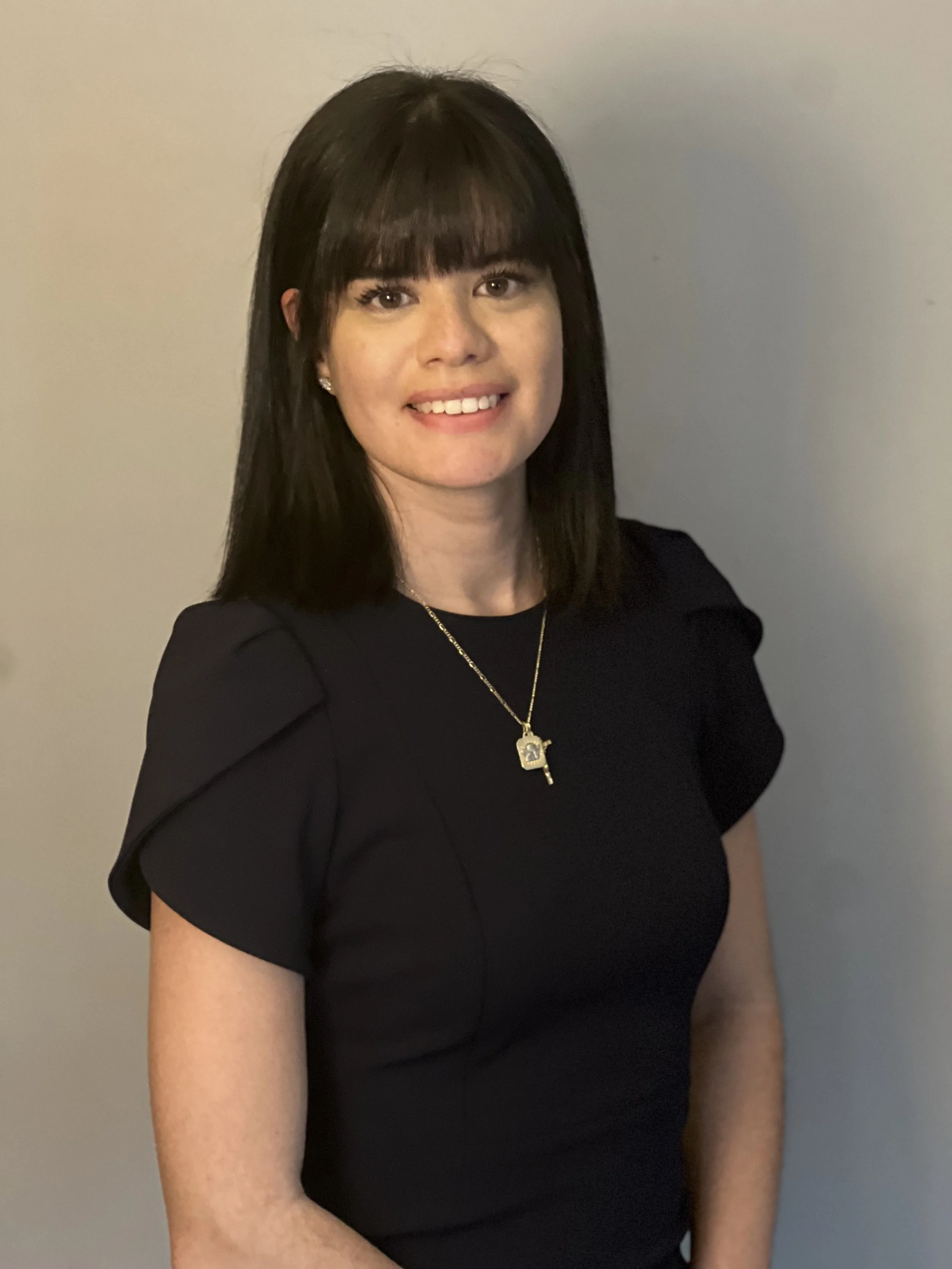 A young woman with straight dark hair and bangs, wearing a black dress with short puffy sleeves, a gold necklace with a house and key pendant, and earrings, smiling against a plain light-colored wall.