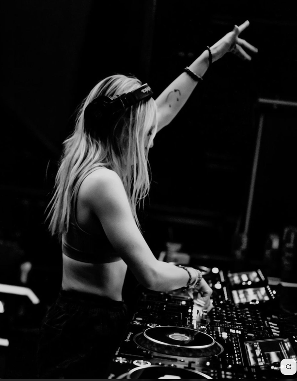 1-on-1 studio DJ Lessons or Music Production Lessons + Daily Zoom Coaching.A female DJ wearing headphones is mixing music at a DJ booth, with one arm raised in the air