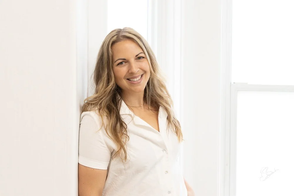 Femme souriante avec cheveux longs, portant une chemise blanche, debout près d'une fenêtre dans une pièce lumineuse.