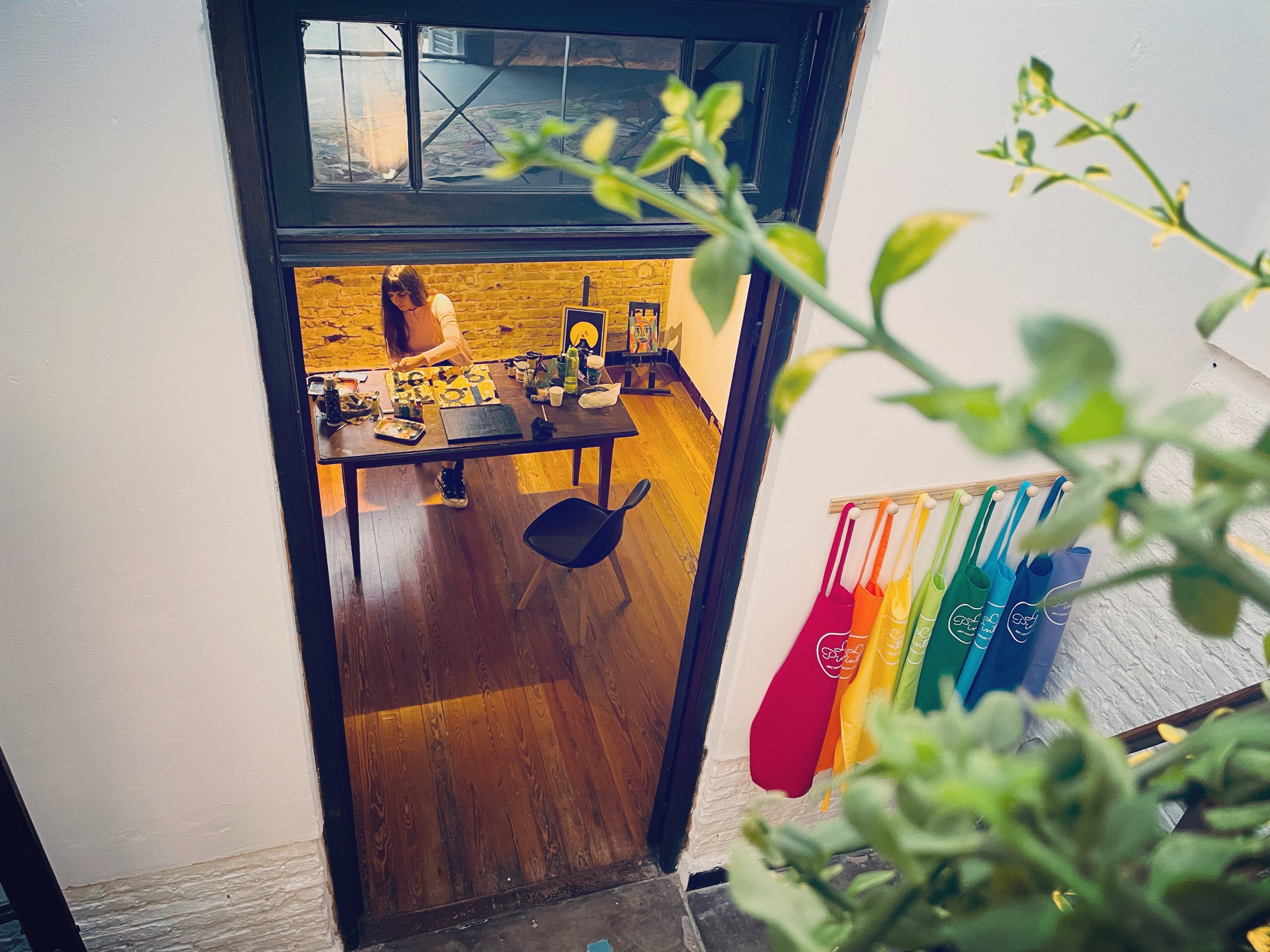 Vista desde arriba de un taller de arte con una persona pintando en una mesa de madera, rodeada de materiales de arte. Hay un conjunto de delantales de colores colgados en la pared al lado de la puerta. En primer plano, se ven hojas verdes de una pla