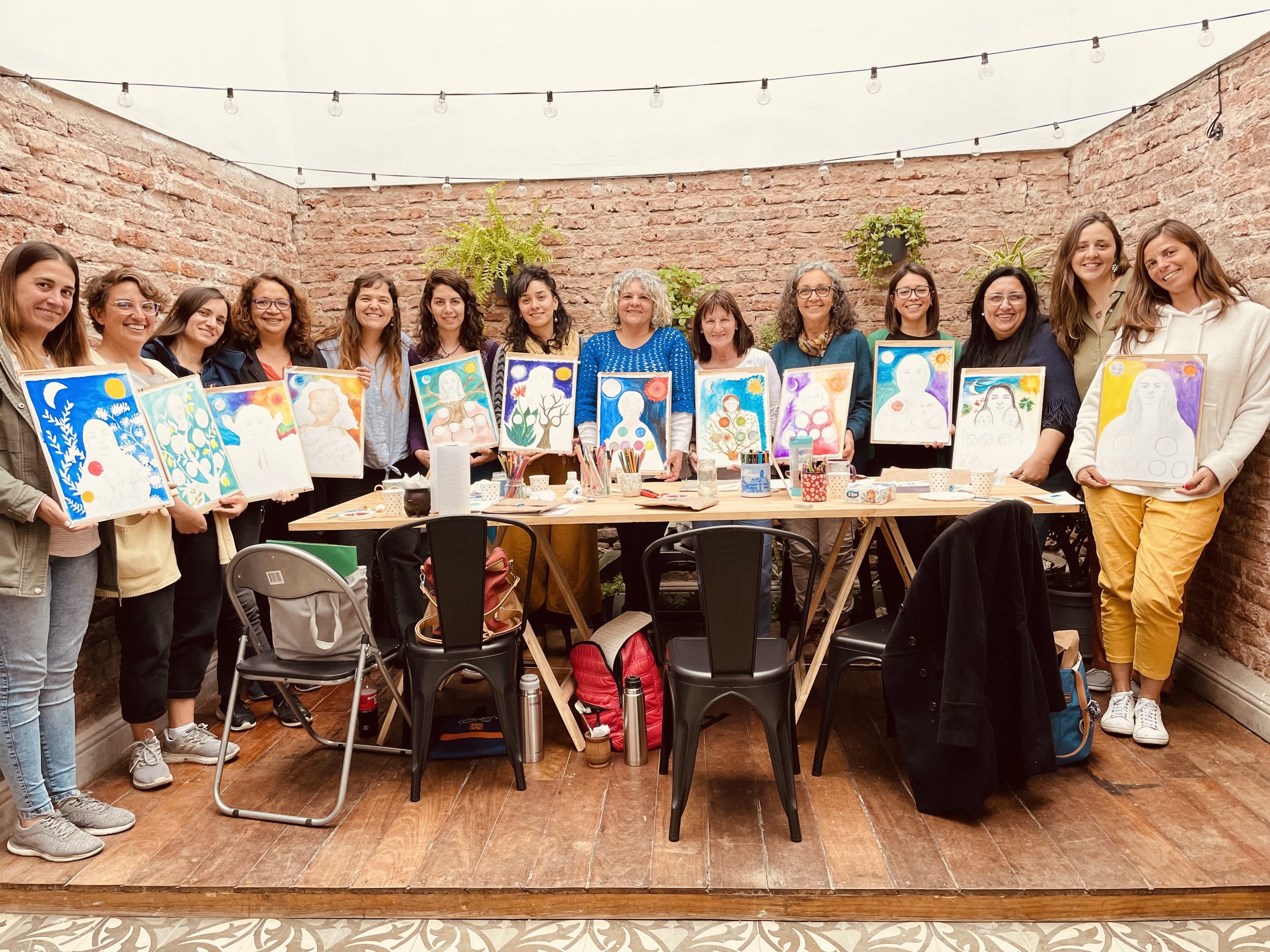 Grupo de mujeres sosteniendo cuadros pintados a mano en un interior con paredes de ladrillo y luces colgantes.