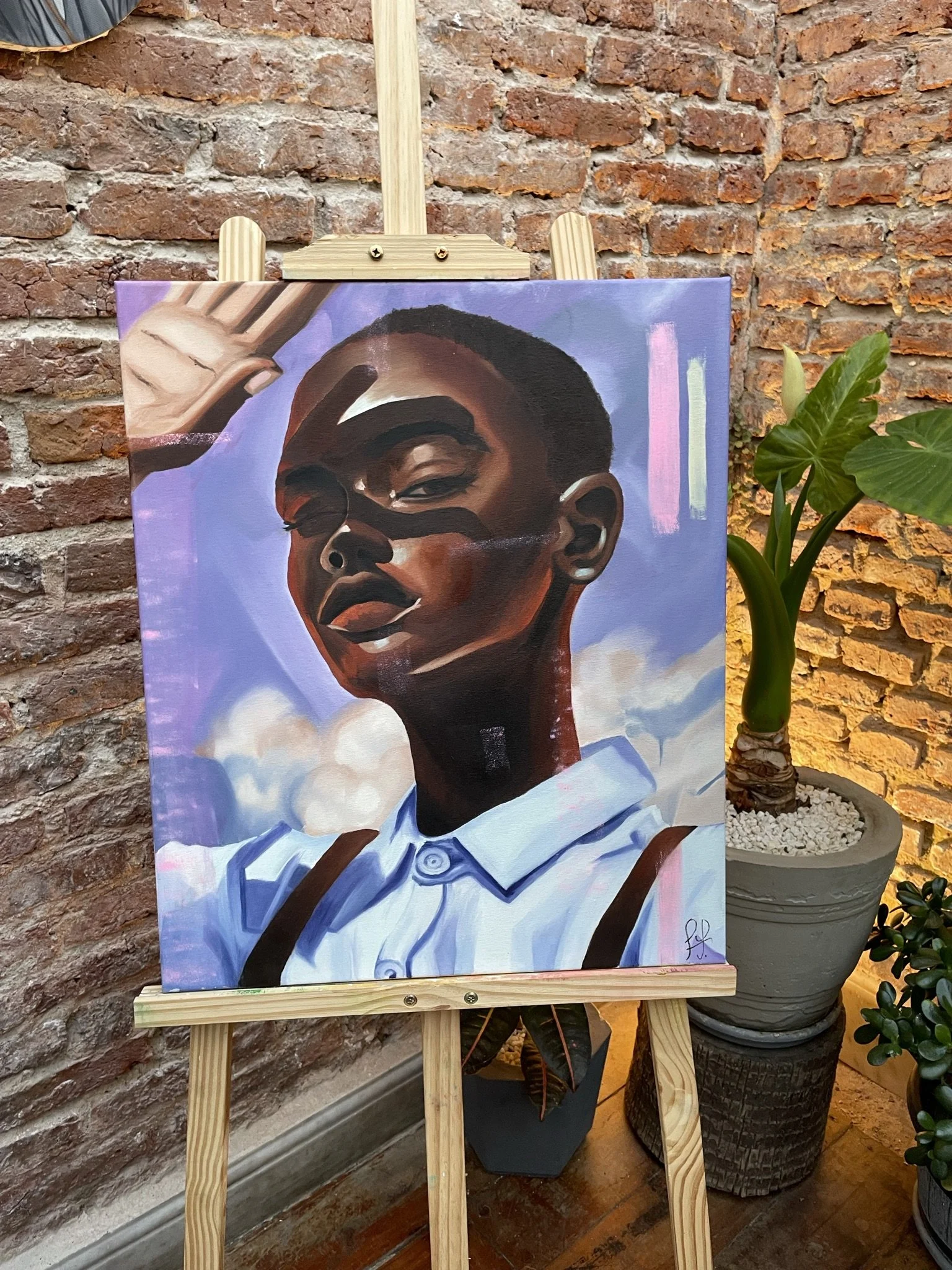 Retrato de una persona afrodescendiente con ojos cerrados y expresión serena, en un cuadro sobre un caballete, con un fondo de ladrillo y plantas decorativas alrededor.