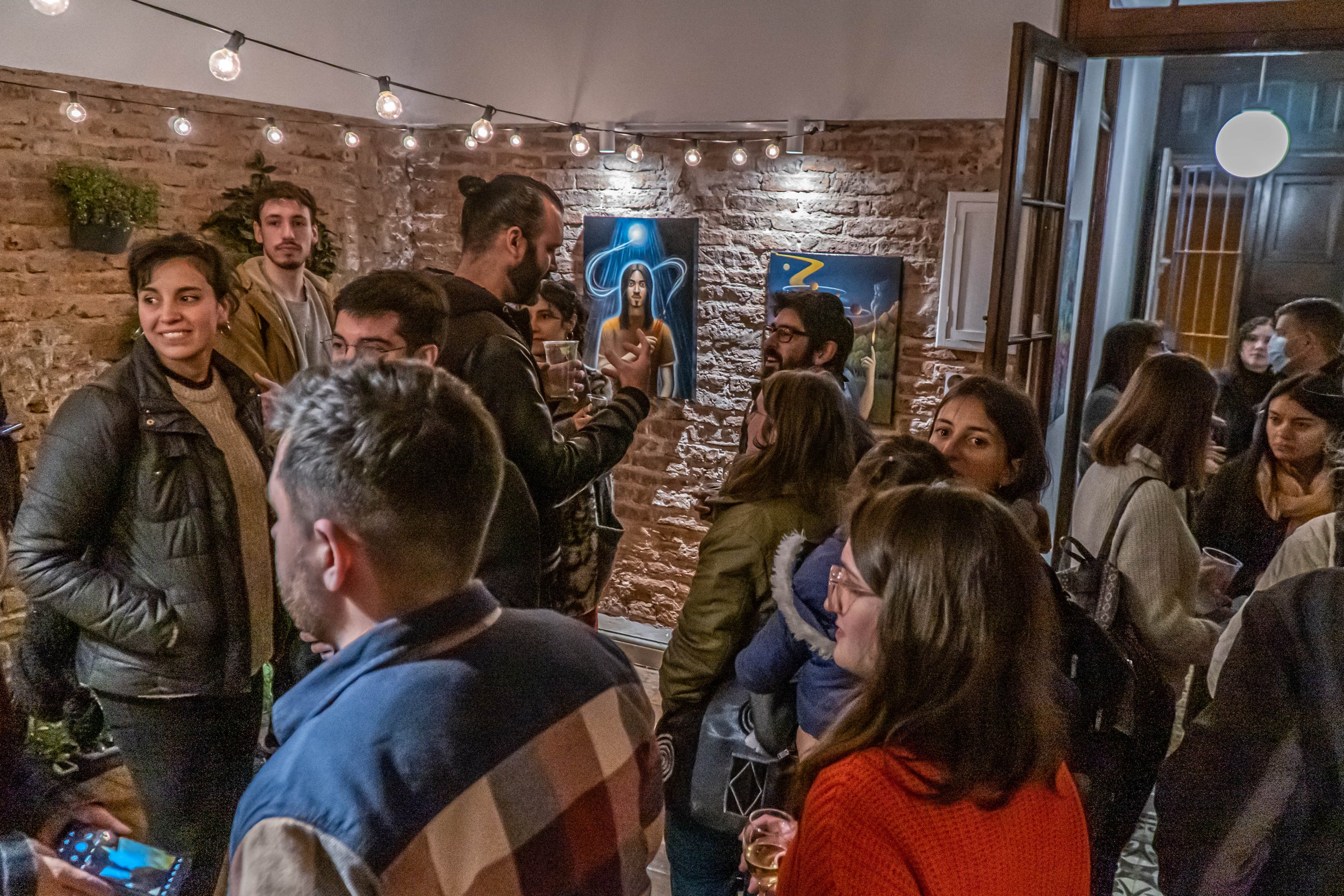 Personas socializando en una galería de arte con luces colgantes y cuadros en la pared.