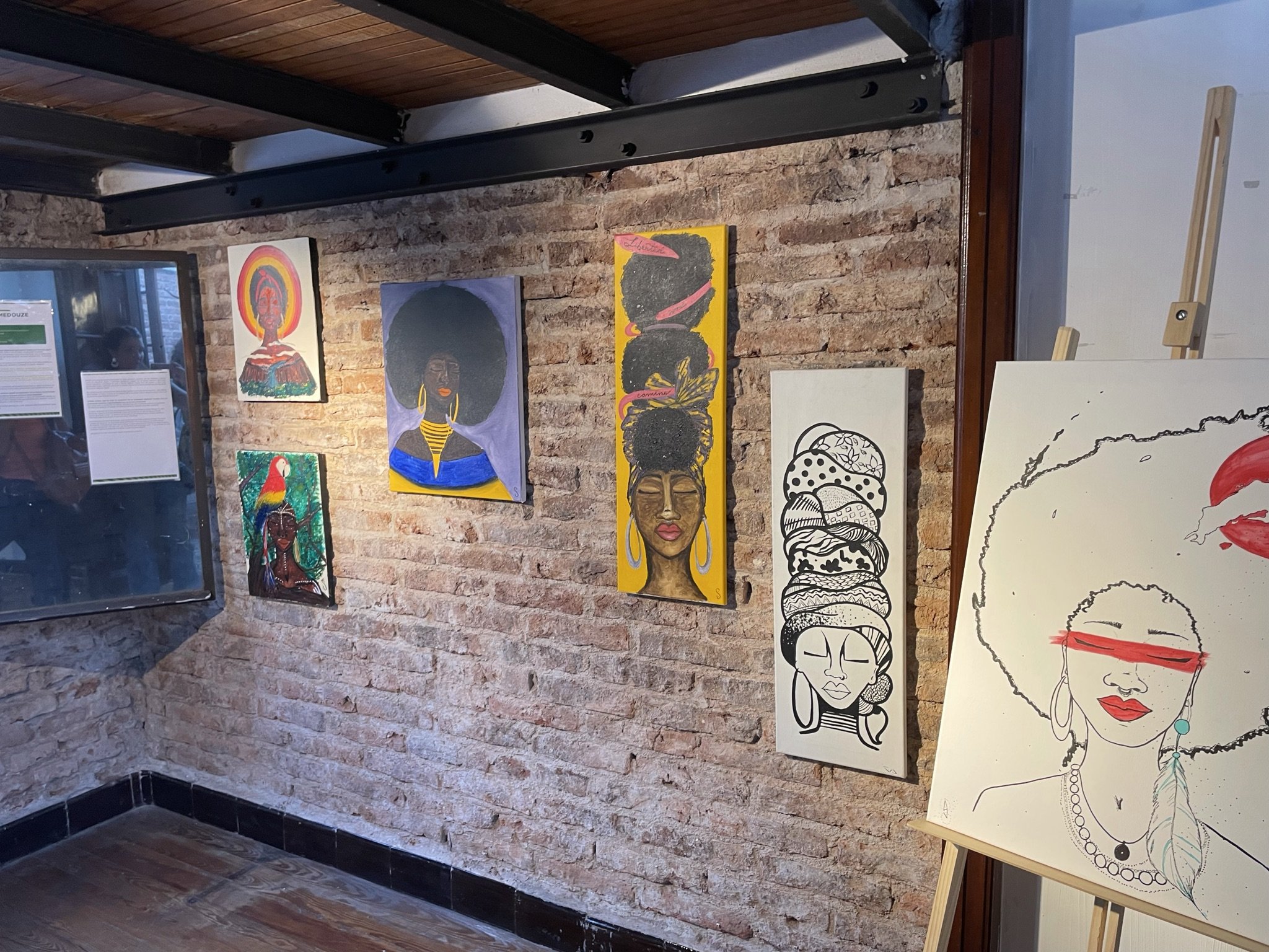 Exposición de arte en una galería con varias pinturas coloridas y retratos de mujeres afrodescendientes en las paredes de ladrillo, junto con un caballete a la derecha.
