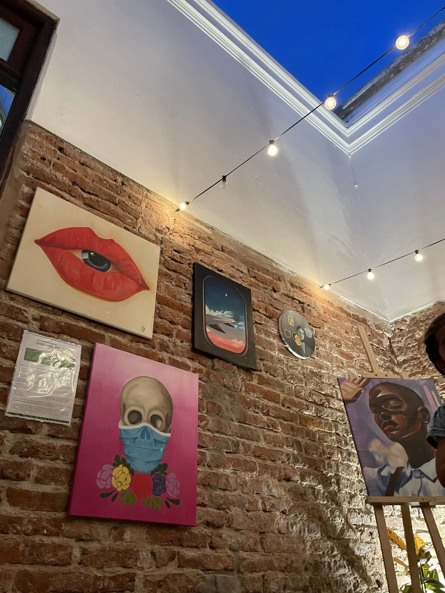 Exhibición de arte en una pared de ladrillo con varios cuadros, incluyendo uno de un ojo y unos labios rojos, una calavera con mascarilla y flores, un paisaje visto desde la ventana de un avión, y un retrato de una persona. Luces colgantes adornan el