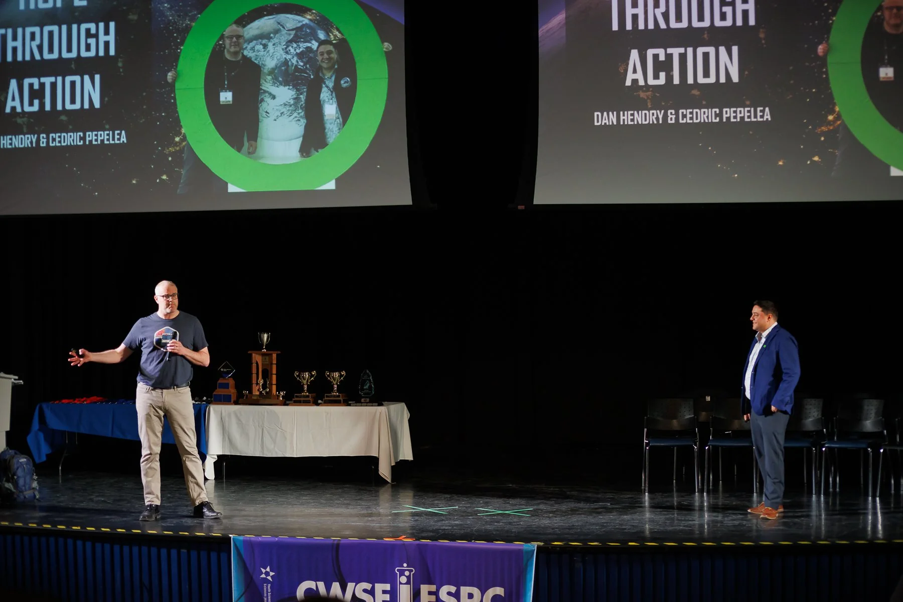 Two presenters on stage during a conference or awards event. The left presenter, a man in a black t-shirt and khaki pants, is speaking and gesturing with his right hand. The right presenter, a man in a blue suit and white shirt, stands facing him. Behind them is a large screen displaying the title 'Through Action' and the names 'Dan Hendry & Cedric Pepelea,' with images of Earth and space. A table on the stage holds several trophies and awards. A banner below the stage reads 'CWISE.'