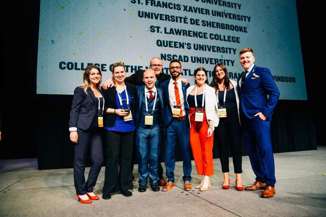 Group of seven young adults wearing business attire, standing arm-in-arm on stage at a college event, with a large screen behind them listing various universities.