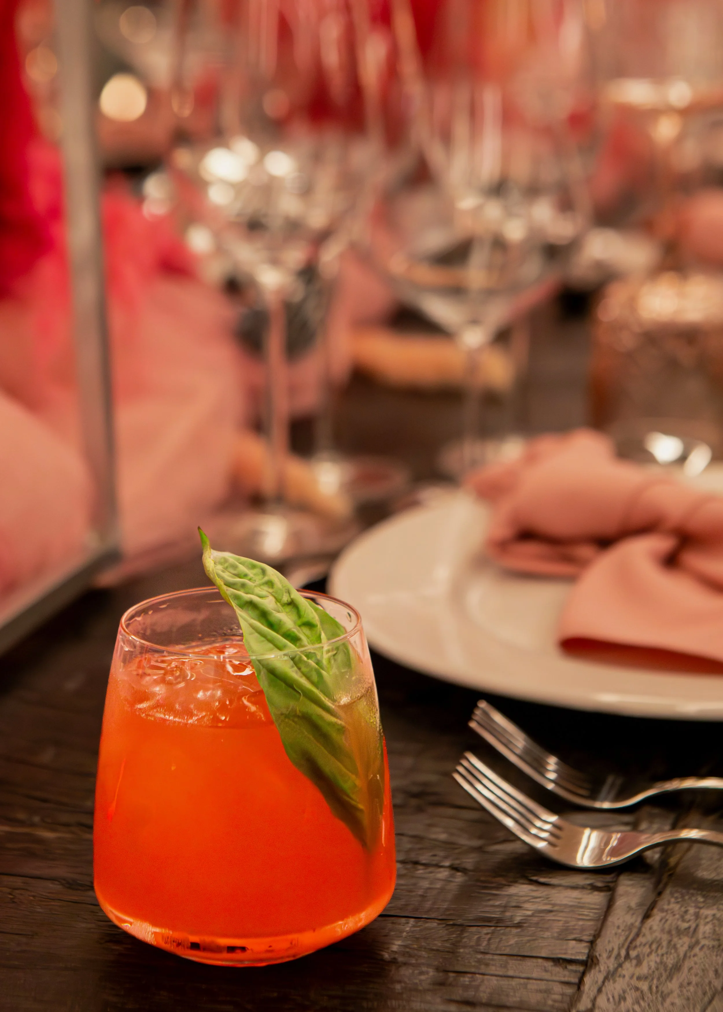 A glass of orange-colored drink with a green basil leaf garnish in the foreground, with a restaurant table setting including plates, silverware, and a blurred background of glasses and pink napkins.