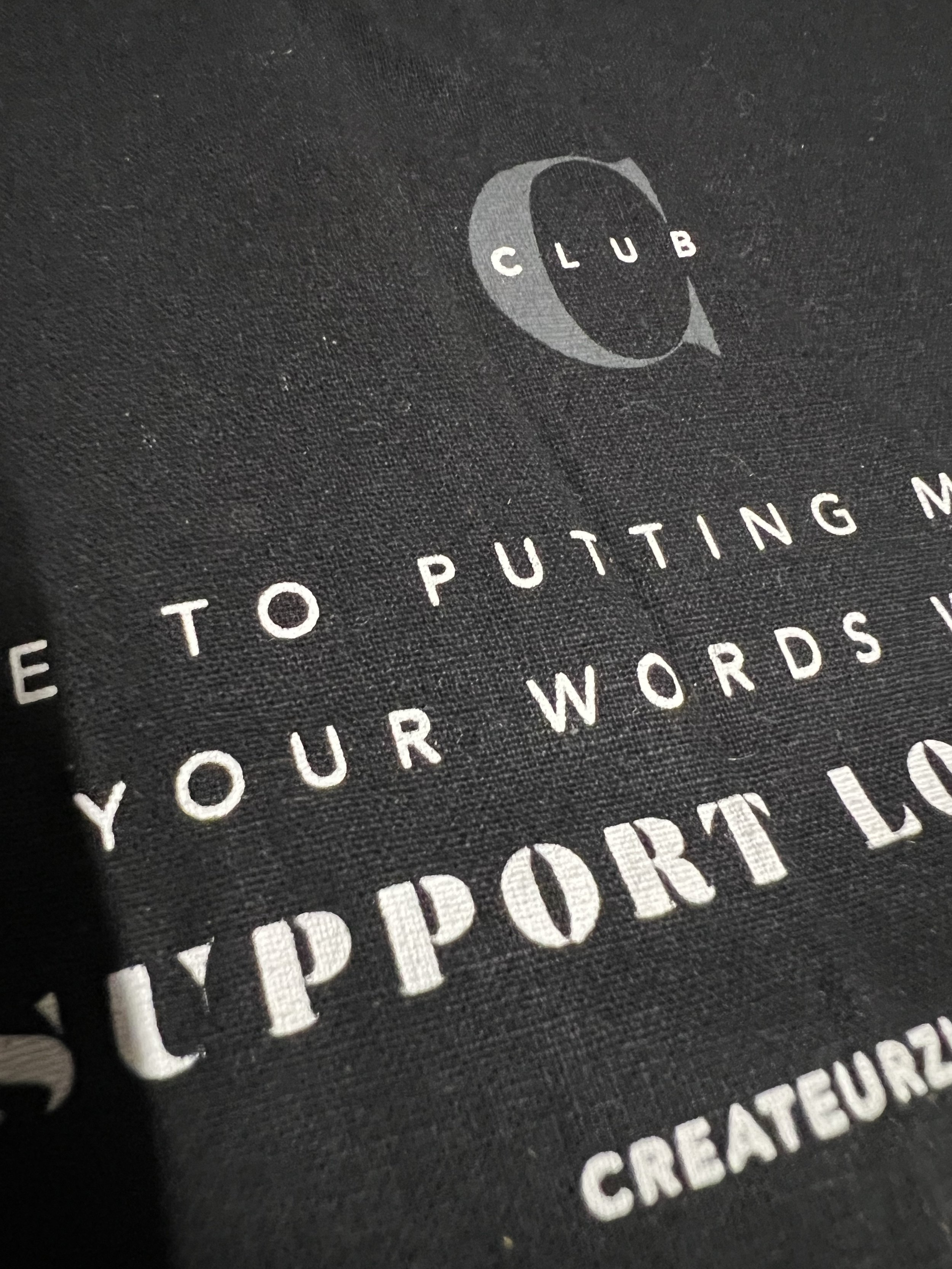 Close-up of a black fabric surface with white text and a logo. The logo is a crescent moon with the word 'CLUB' inside it. The text reads 'HOW TO PUTTING YOUR WORDS SUPPORT LOCAL BUSINESS and 'CREATORE' at the bottom.