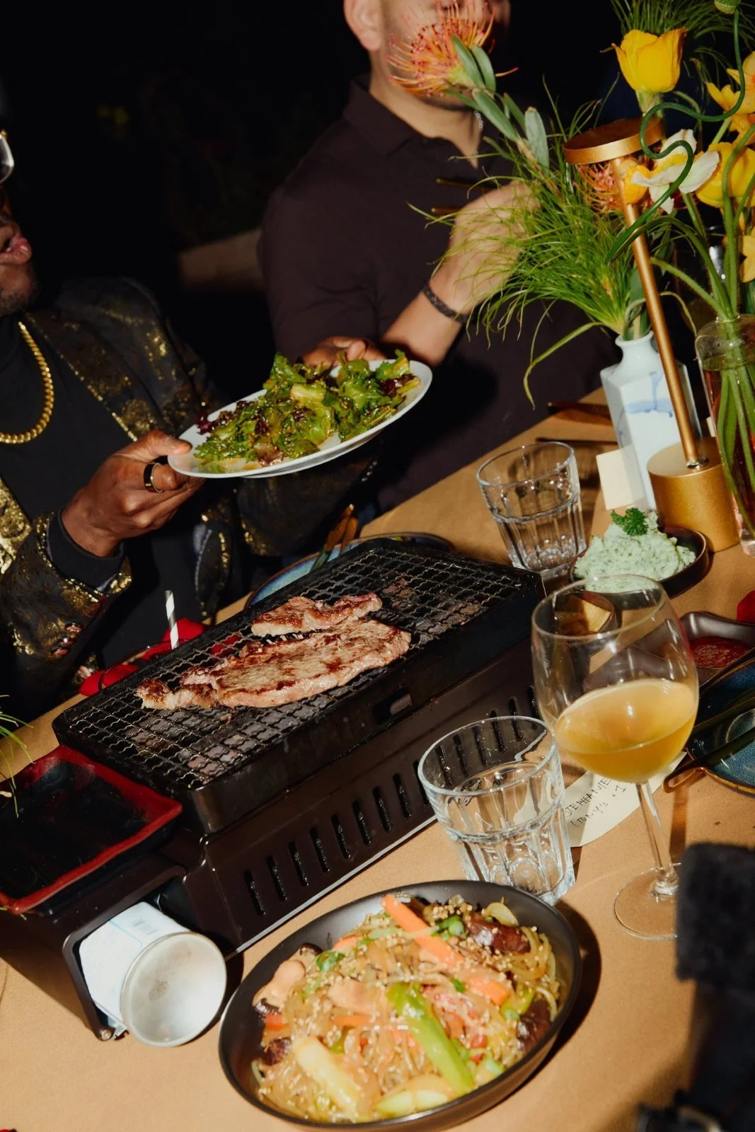 Guests sharing a tabletop Korean BBQ dinner with steak on the grill and japchae