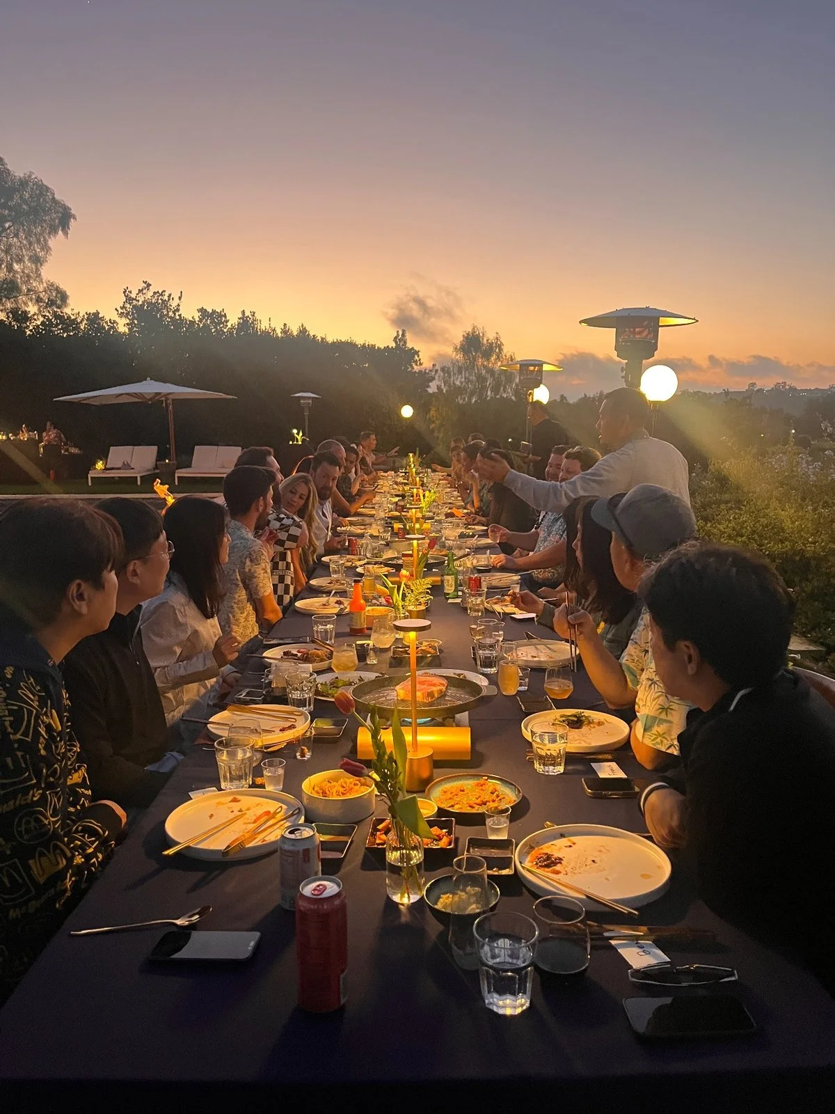 Guests at a long outdoor Korean BBQ dinner table at sunset