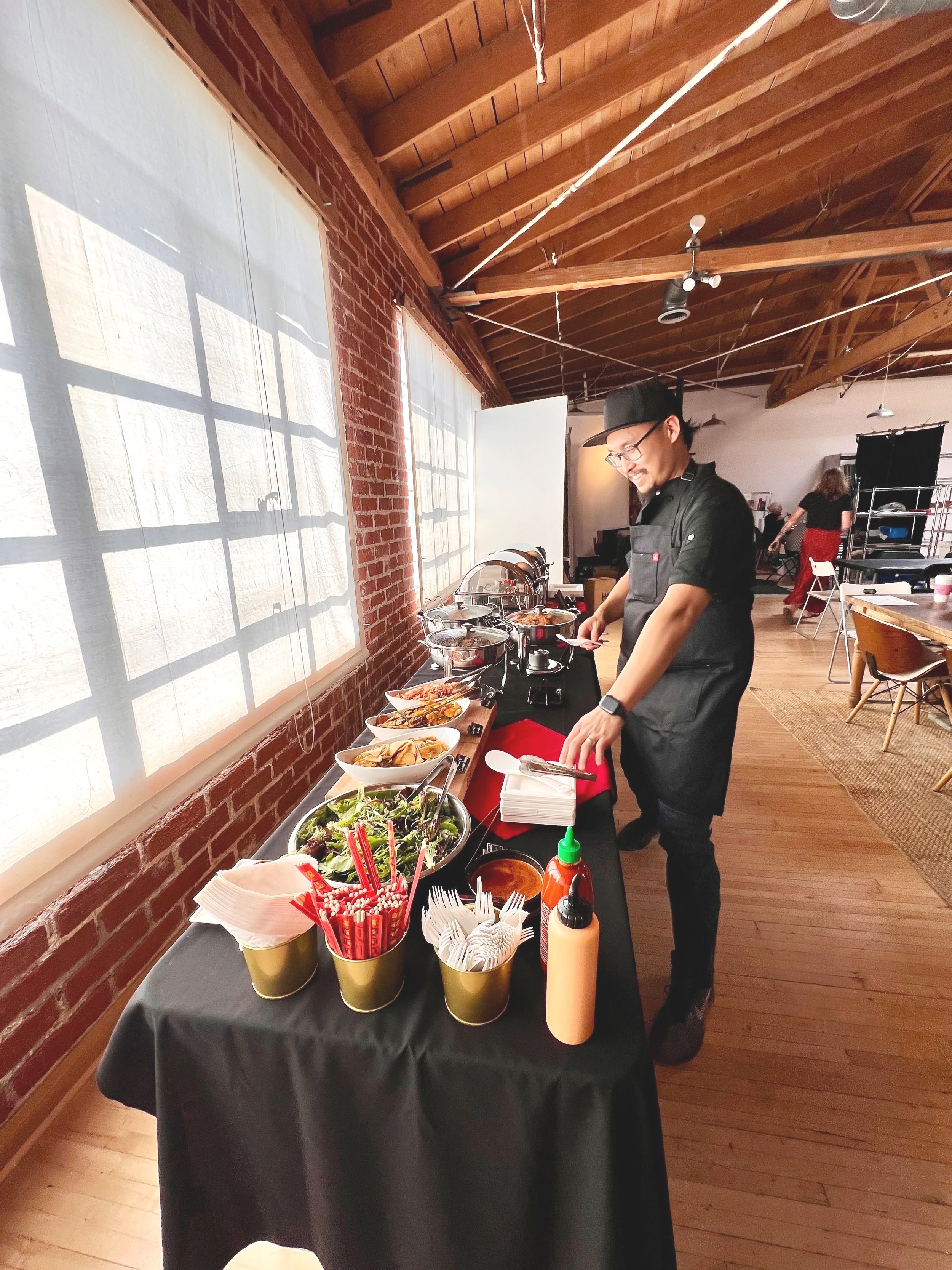 A chef in black uniform and glasses setting up a buffet table with salads, condiments, and dishes in a bright, rustic restaurant with wooden beams and large windows.