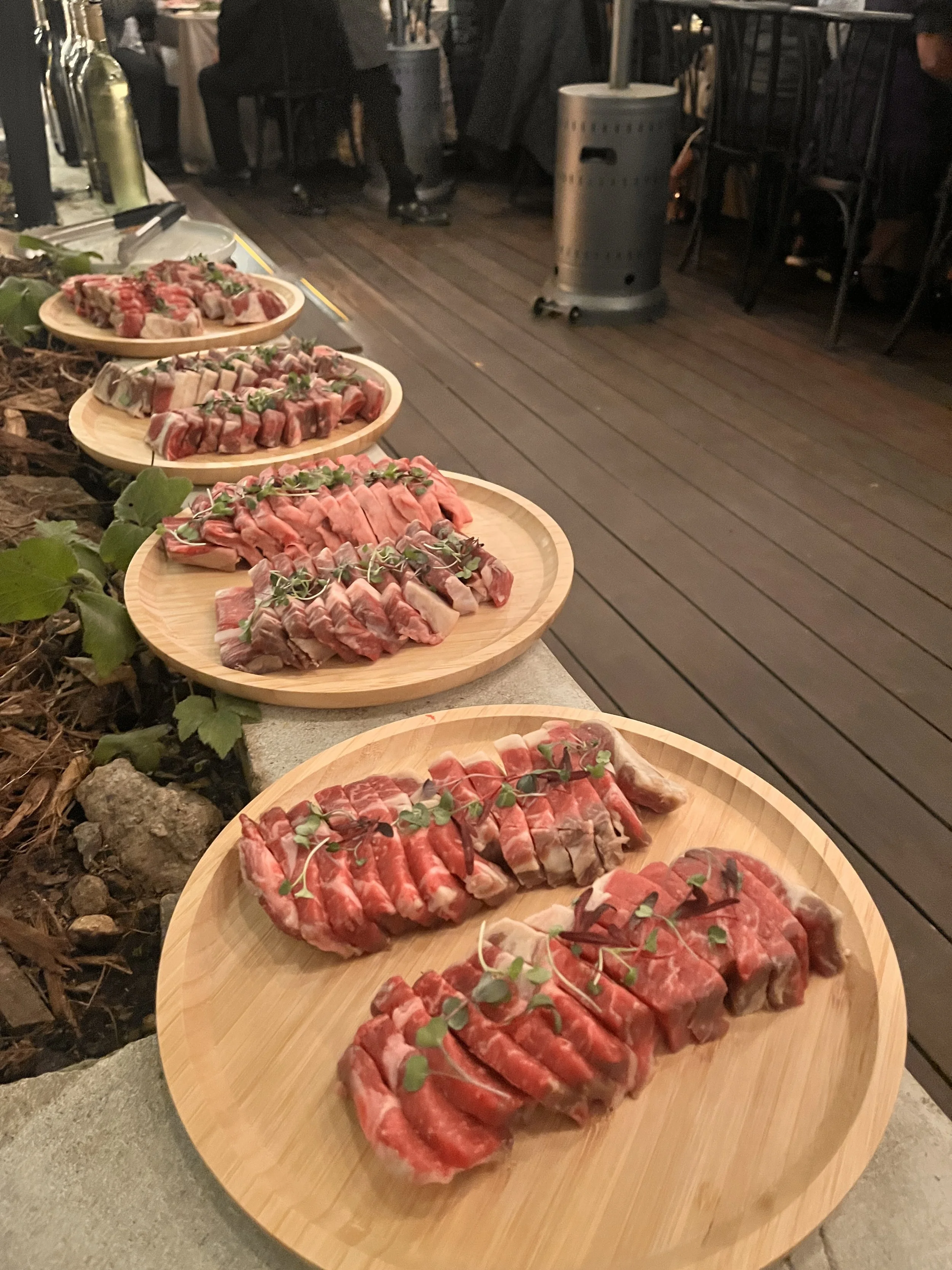 Several wooden platters with raw, seasoned meat garnished with green herbs, arranged on a table outside at night prepared for grilling on table-top grill for Korean Barbecue dinner.
