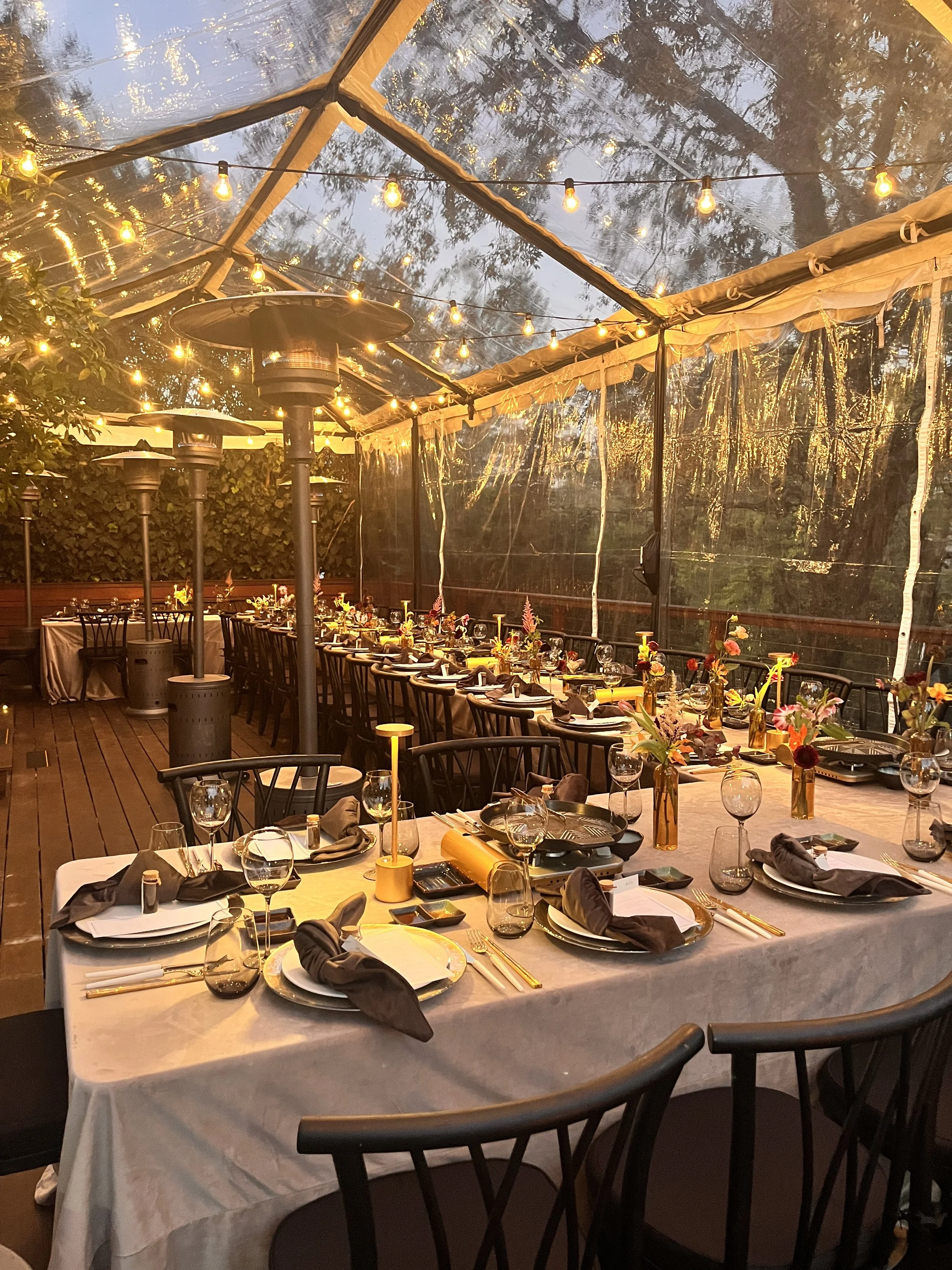 Decorated outdoor dining area with long tables covered in white tablecloths, set with table-top grills for Korean Barbecue, wine glasses, black napkins, and plates. 