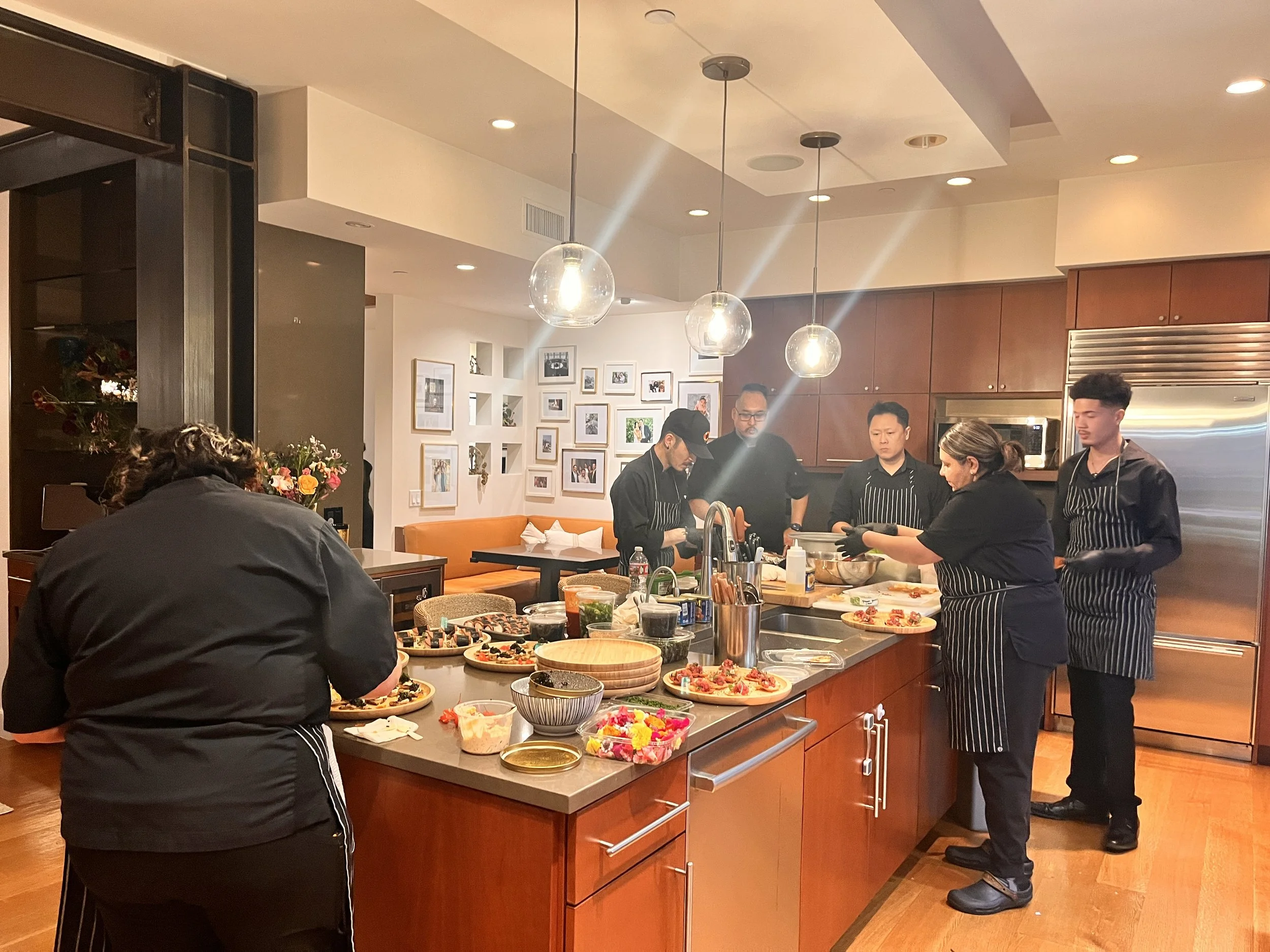 Catering stuff are cooking and preparing food in a modern kitchen with wooden cabinets, a marble countertop, and pendant lights hanging from the ceiling.