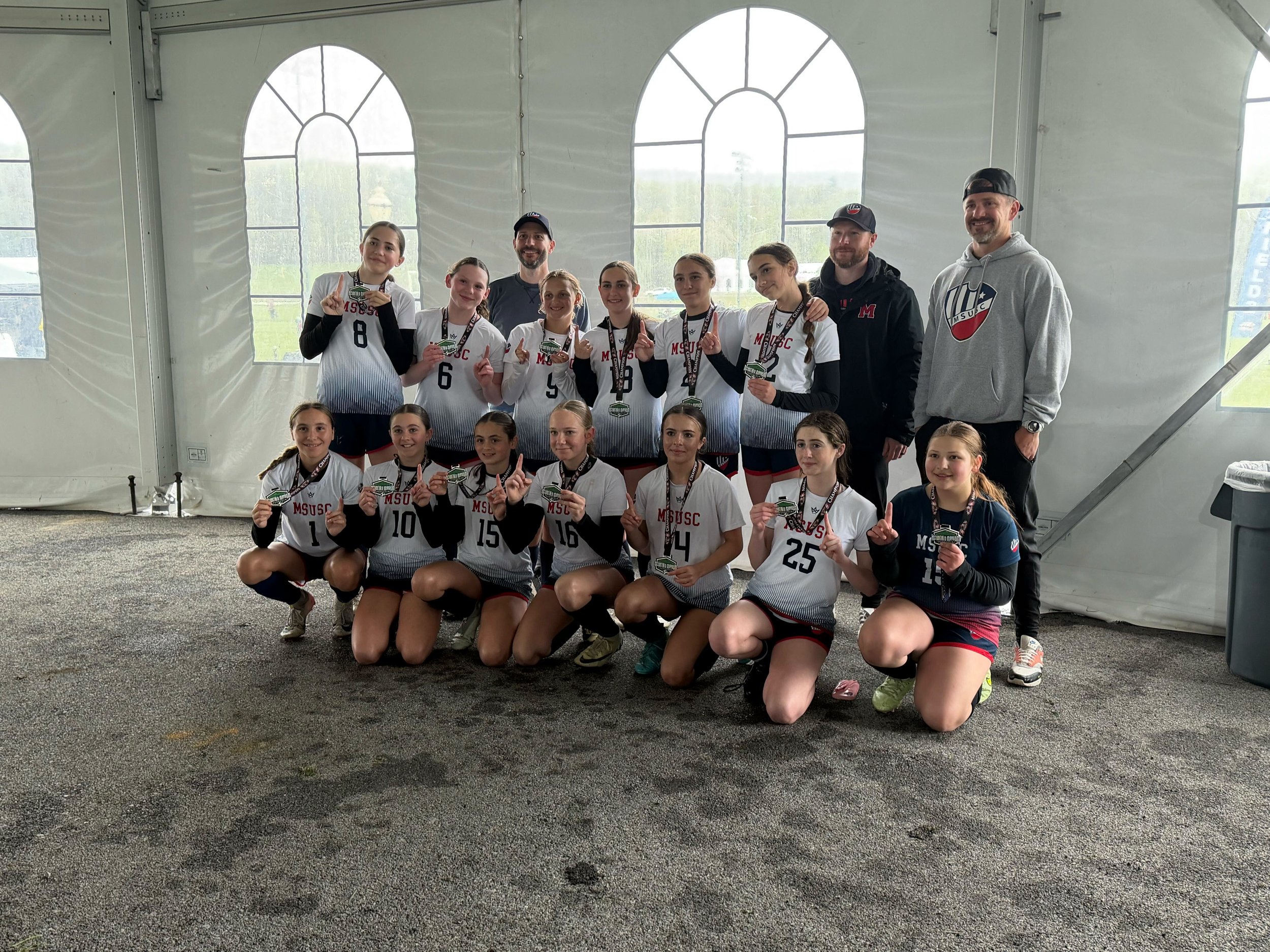 Group of young female volleyball players wearing white and black uniforms with medals, posing with their coaches inside a large tent with arched windows, celebrating their victory.