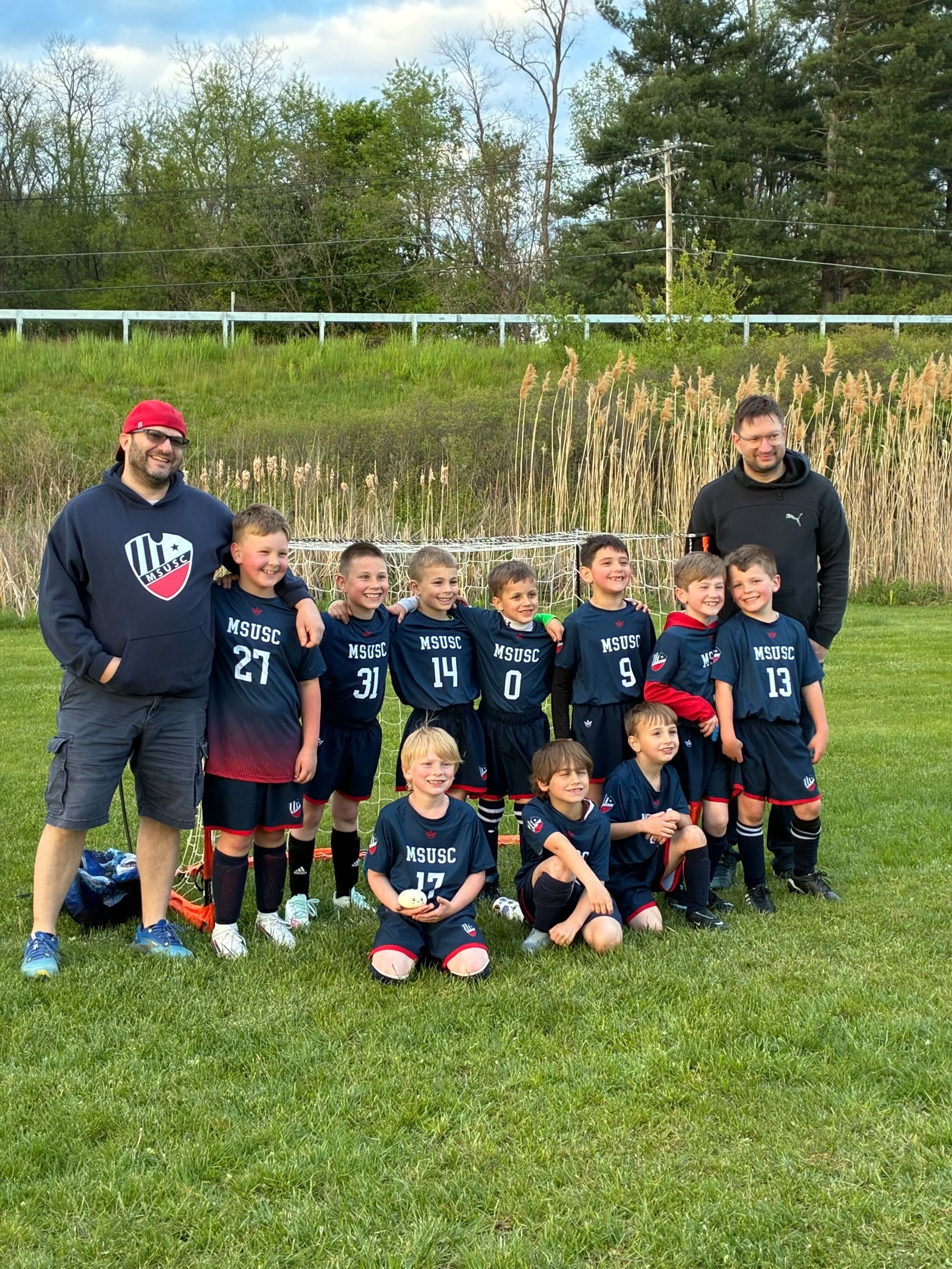 A youth soccer team with two coaches poses on a field after a game. The boys are wearing navy blue jerseys, some with numbers and 'MSUSC' printed on them. The coaches are standing at either end, one in a black hoodie and glasses, the other in a navy 