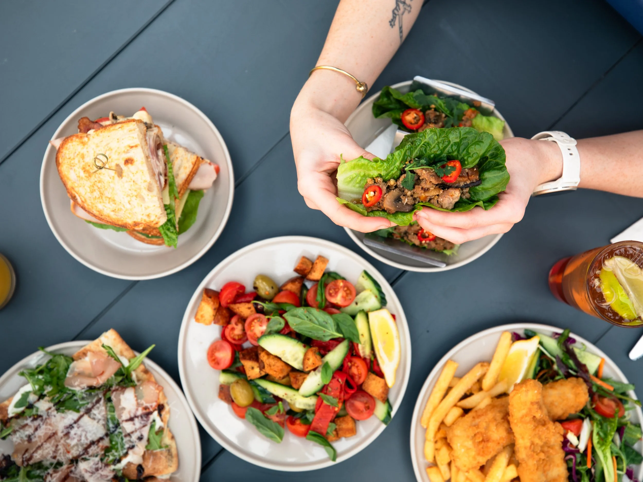 A top down photo of a  person sitting at a table holding a san choy bow with fish and chips, salads, flatbreads and club sandwiches across the table