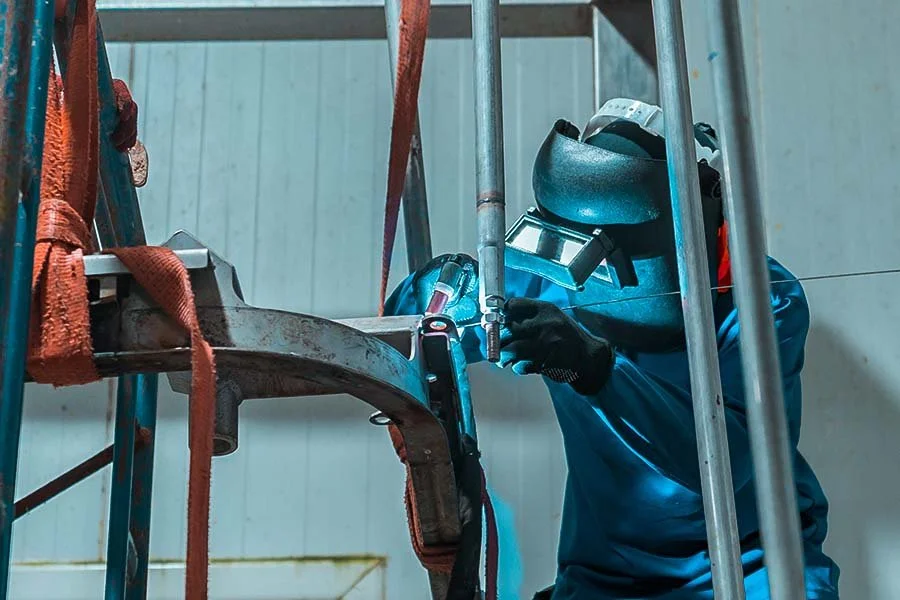 A worker welding metal pipes on a scaffold in a workshop, wearing a welding helmet and protective gloves.