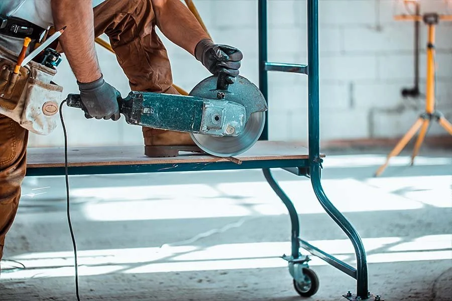 A worker cutting a piece of wood with a circular saw on a metal work cart in a workshop or construction site.