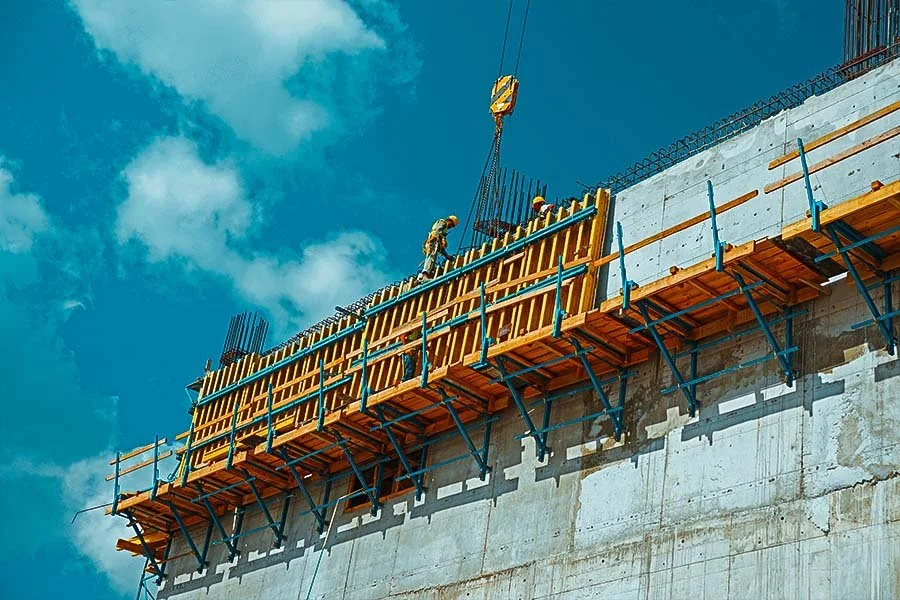 Construction workers building a bridge or elevated structure, with blue sky and clouds in the background.