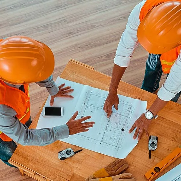 Two construction workers wearing orange safety vests and helmets reviewing blueprints on a wooden table.