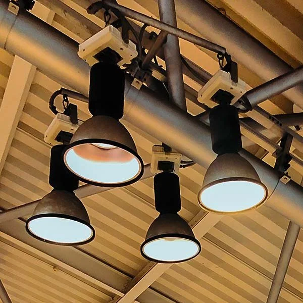 Four large industrial ceiling lights turned on in a building, attached to a metal pipe ceiling framework.