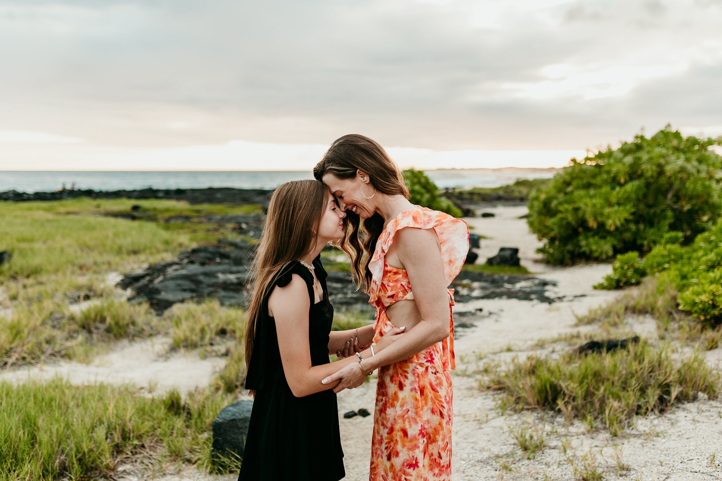 A woman and girl stand close together on a sandy beach, touching foreheads and smiling, with ocean and trees in the background during sunset.