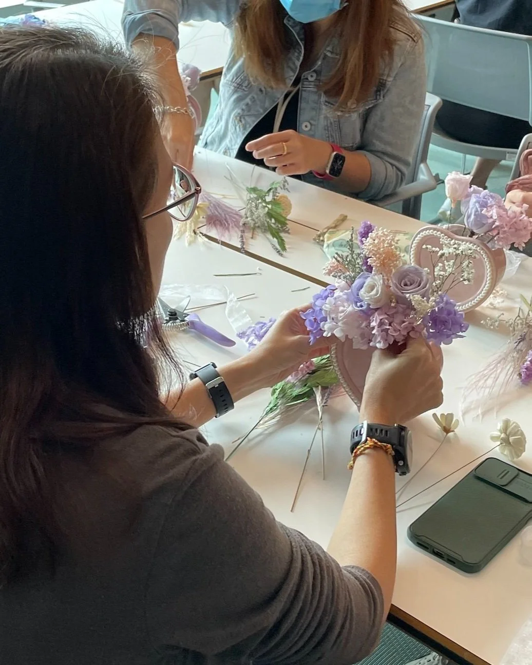 A woman with glasses and a watch is arranging purple, white, and pink flowers in a floral headband, seated at a table with crafting materials, other flowers, and a smartphone nearby.