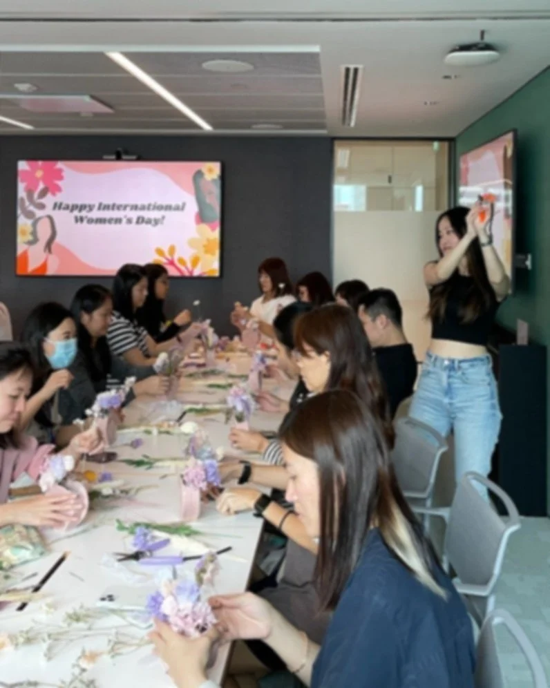 Group of women sitting at a long table with floral decorations, participating in a Women's Day celebration, with a woman taking a photo and a screen displaying "Happy International Women's Day!" in the background.