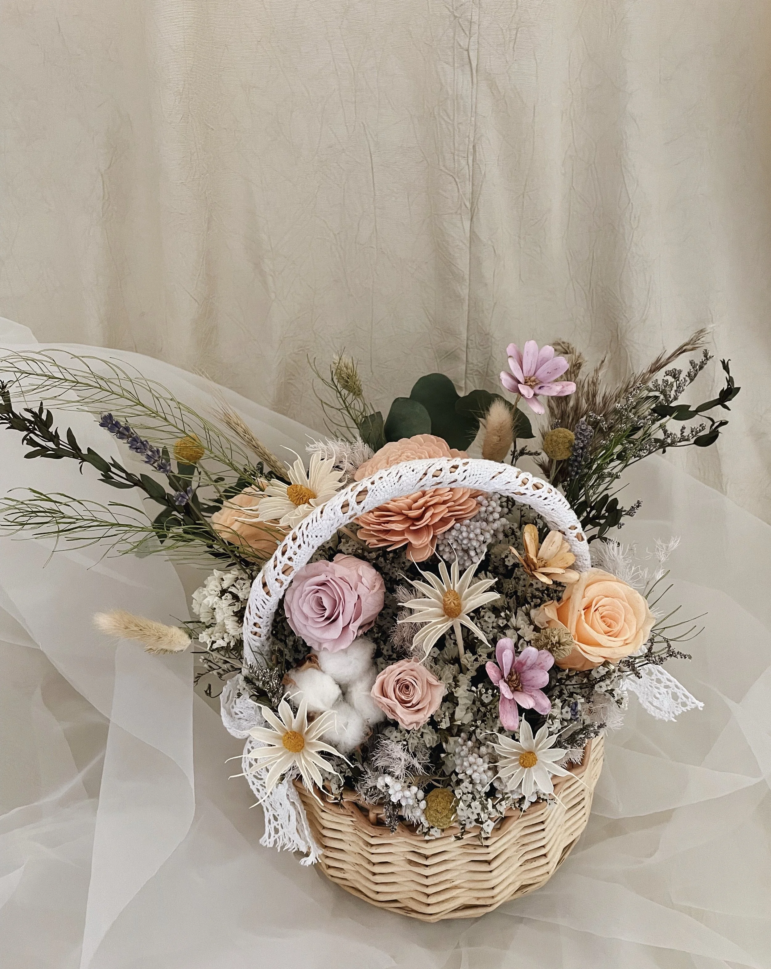 A wicker basket filled with a variety of pink, white, and peach flowers, including roses, daisies, and other blooms, arranged with greenery and decorative accents, set against a neutral background.