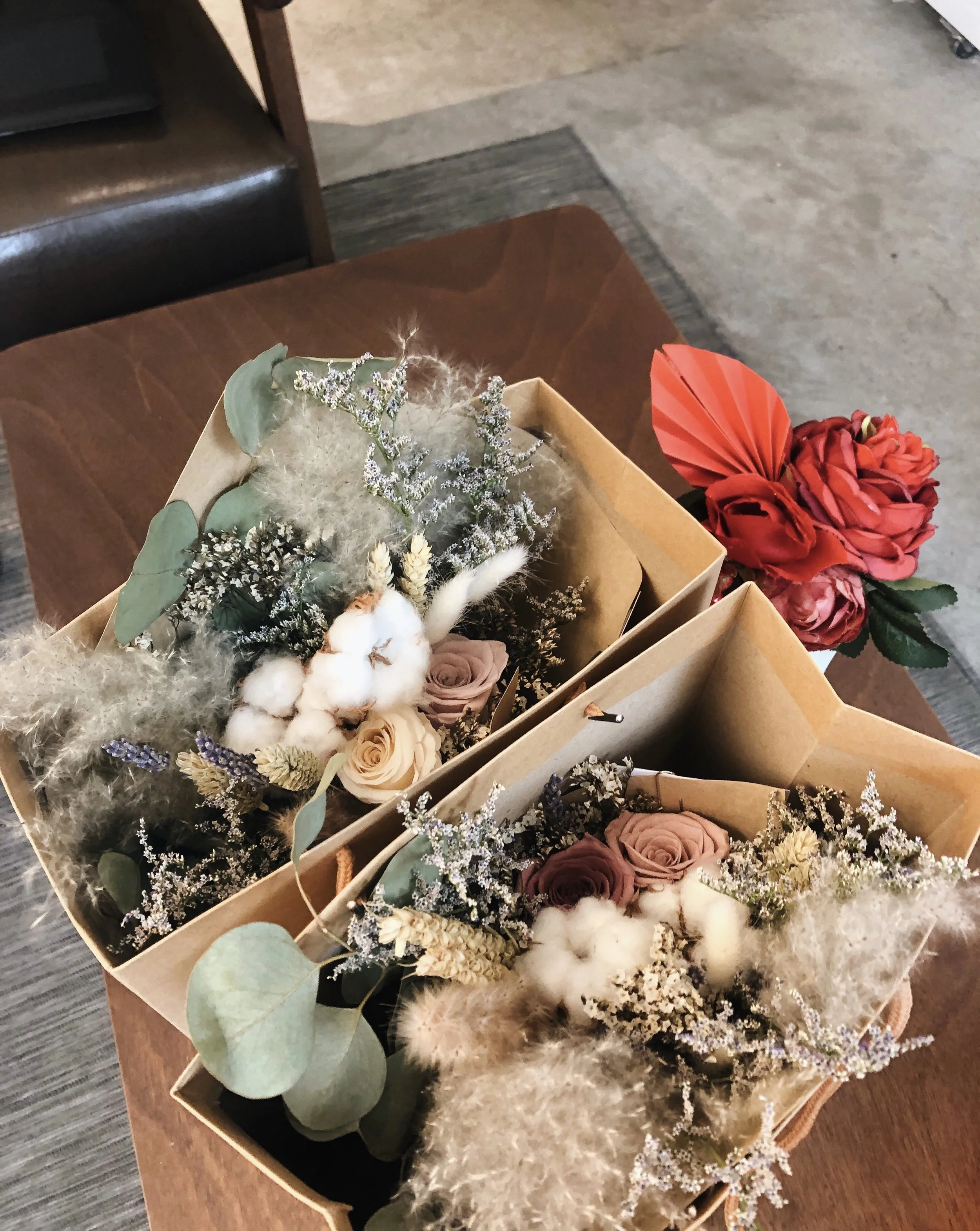 Various floral bouquets featuring roses, dried flowers, eucalyptus, and cotton, arranged in brown paper wrapping on a wooden table.