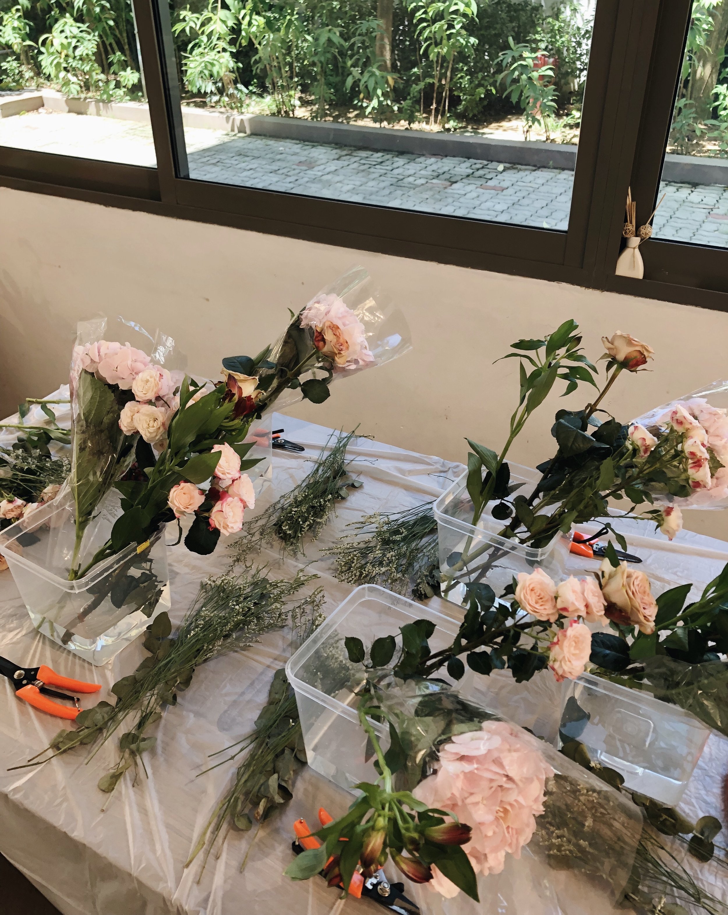 A table with various flowers and greenery arranged for floral arrangements, with scissors and plastic containers, next to a window with greenery outside.