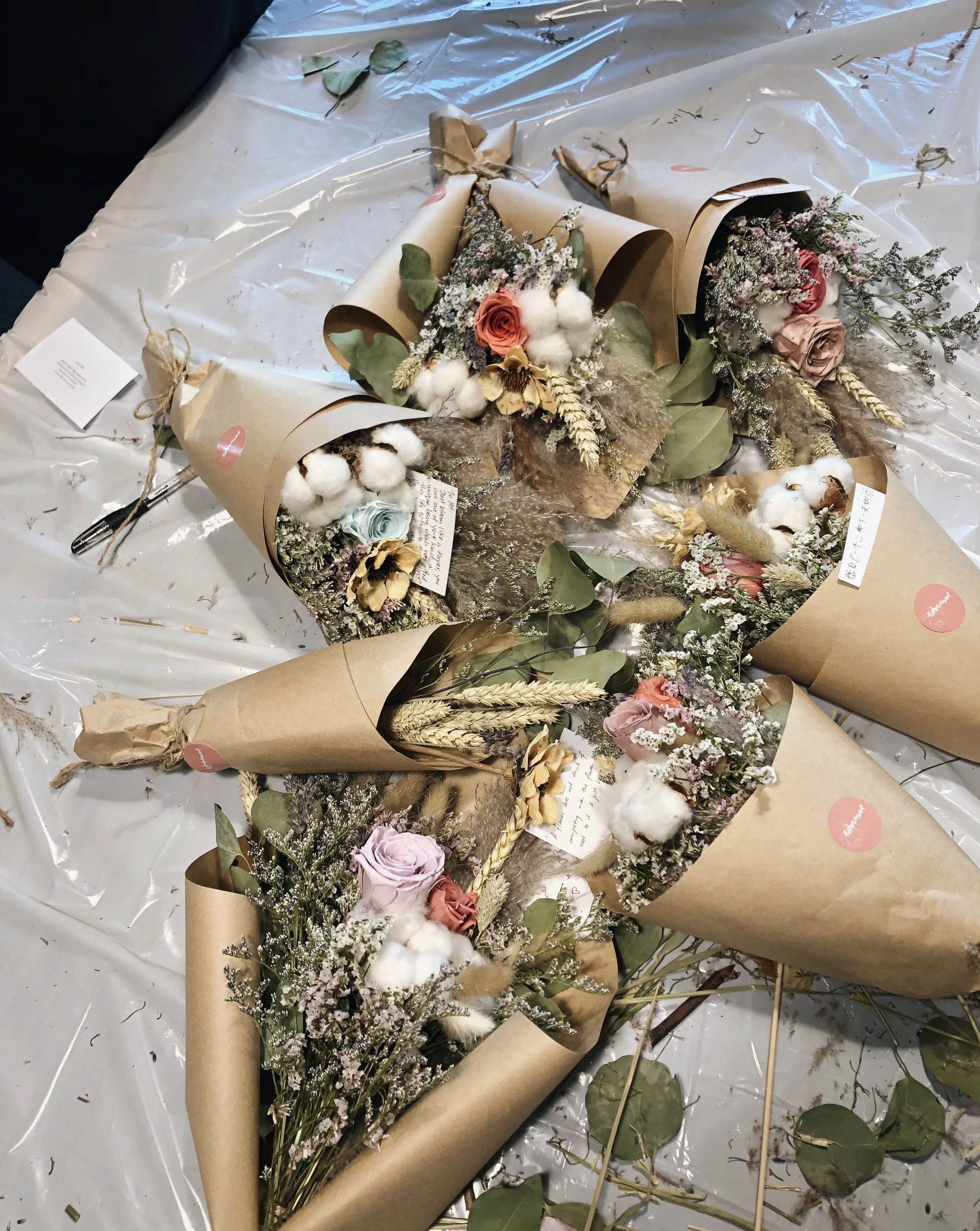 Multiple bouquets of flowers wrapped in brown craft paper, arranged on a plastic-covered surface, with some leaves and flower stems scattered around.