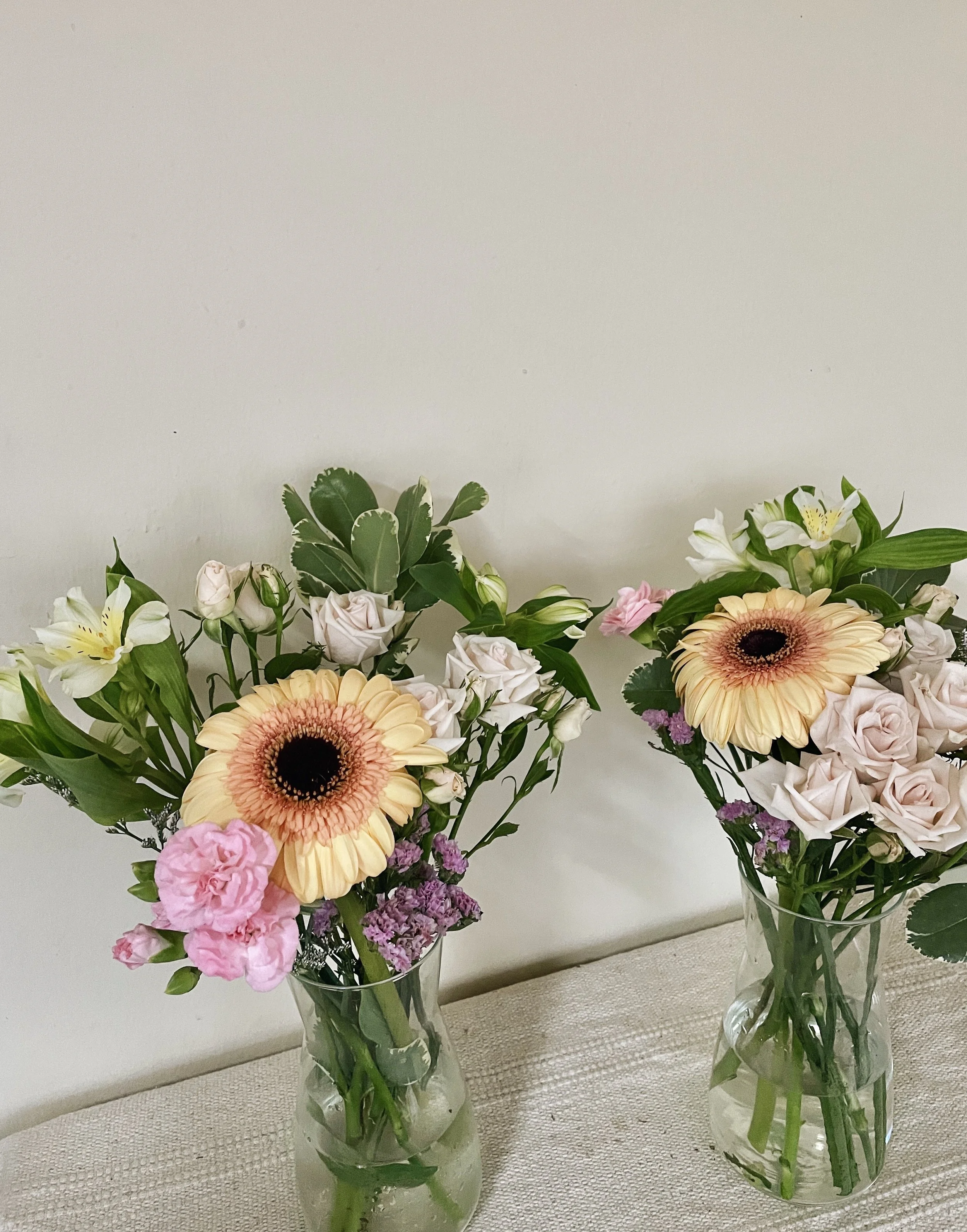 Two glass vases with assorted fresh flowers, including gerbera daisies, roses, and lilies, placed on a table against a light-colored wall.