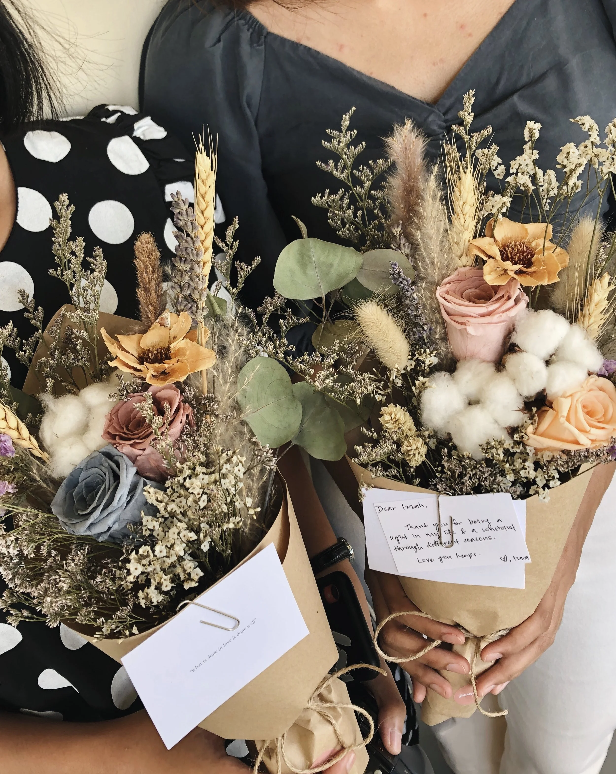 Two women holding bouquets of mixed flowers, including roses, dried wheat, eucalyptus, and cotton, with handwritten notes attached.