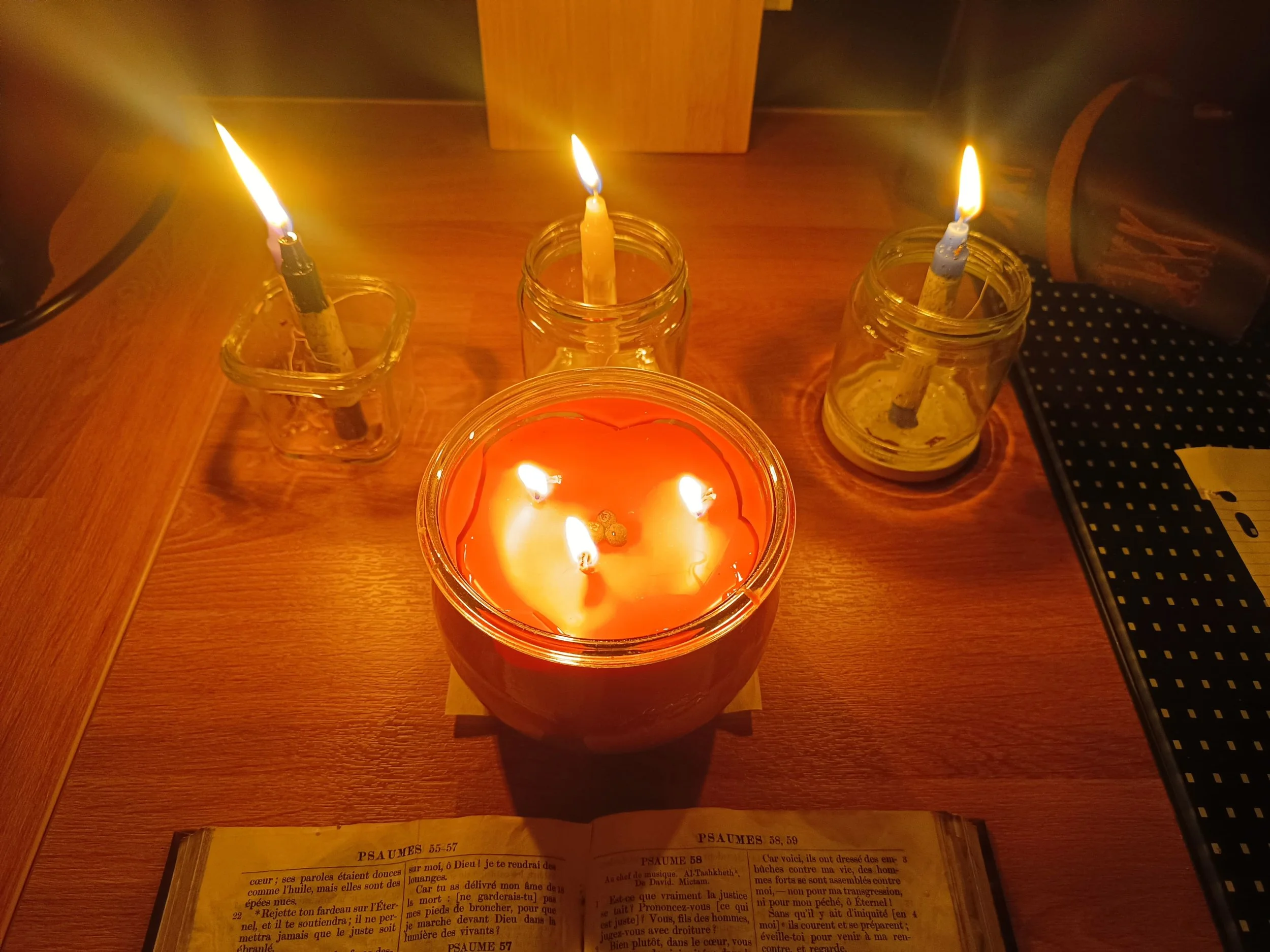 Trois bougies allumées dans des petits pots en verre sur une table en bois, une bougie orange à l'avant, deux autres derrière. Il y a aussi un livre ouvert avec des psaumes en dessous.
