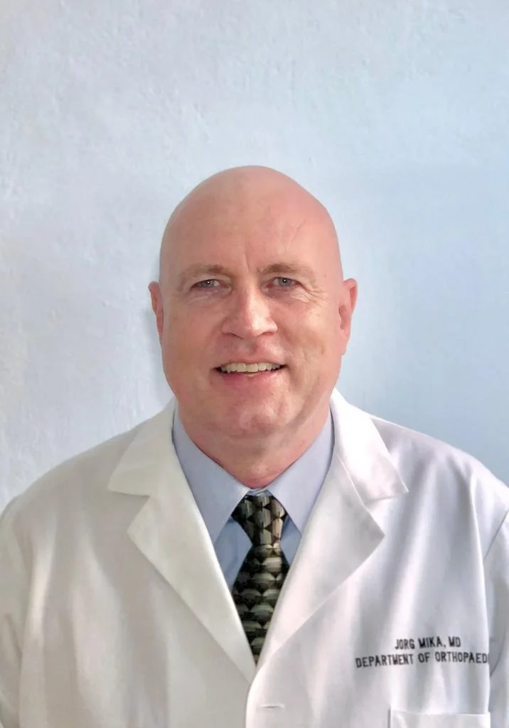 A man with a shaved head and blue eyes wearing a dark striped suit, white shirt, and dark tie, looking directly at the camera, against a plain light background.